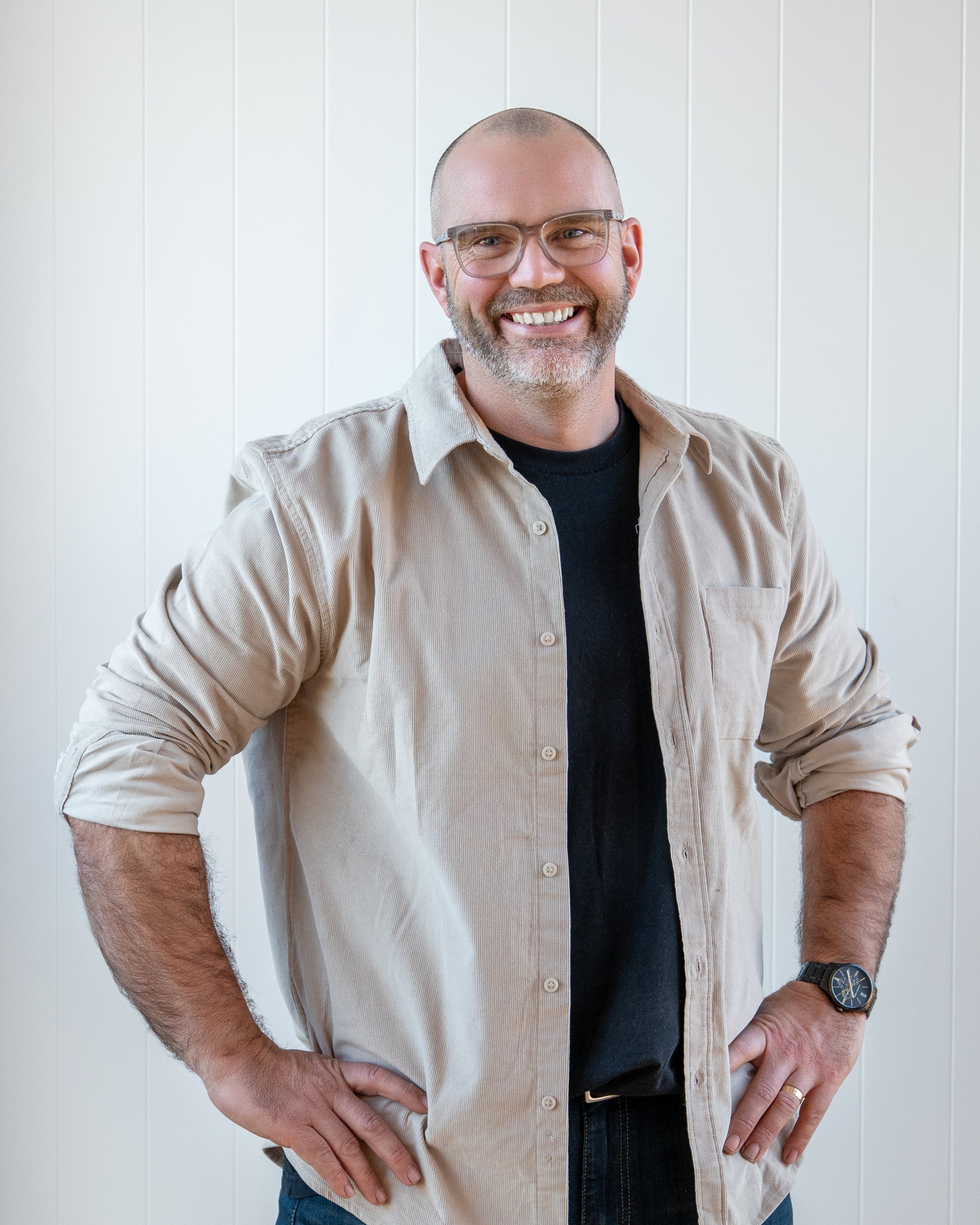 A smiling man with glasses, a beard, and a shaved head, wearing a beige button-up shirt over a black t-shirt, standing with hands on hips in front of a white wall. Renovations, Extensions & Additional Dwellings Canterbury | Alterhouse