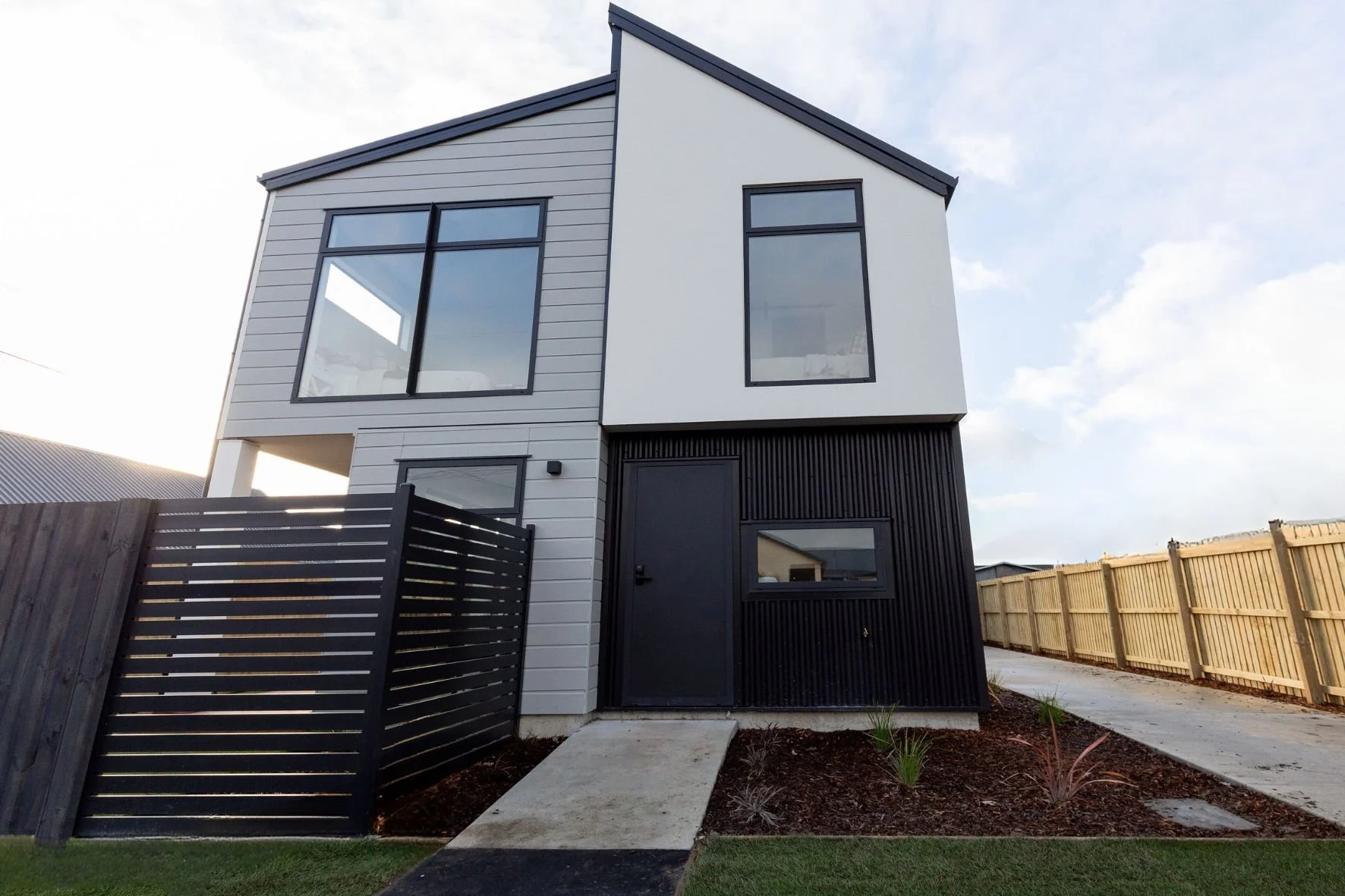 Modern two-story house with black and white exterior, large windows, black fence and gate, and a pathway leading to the front door, with a wooden fence on the right side and a partly cloudy sky in the background.