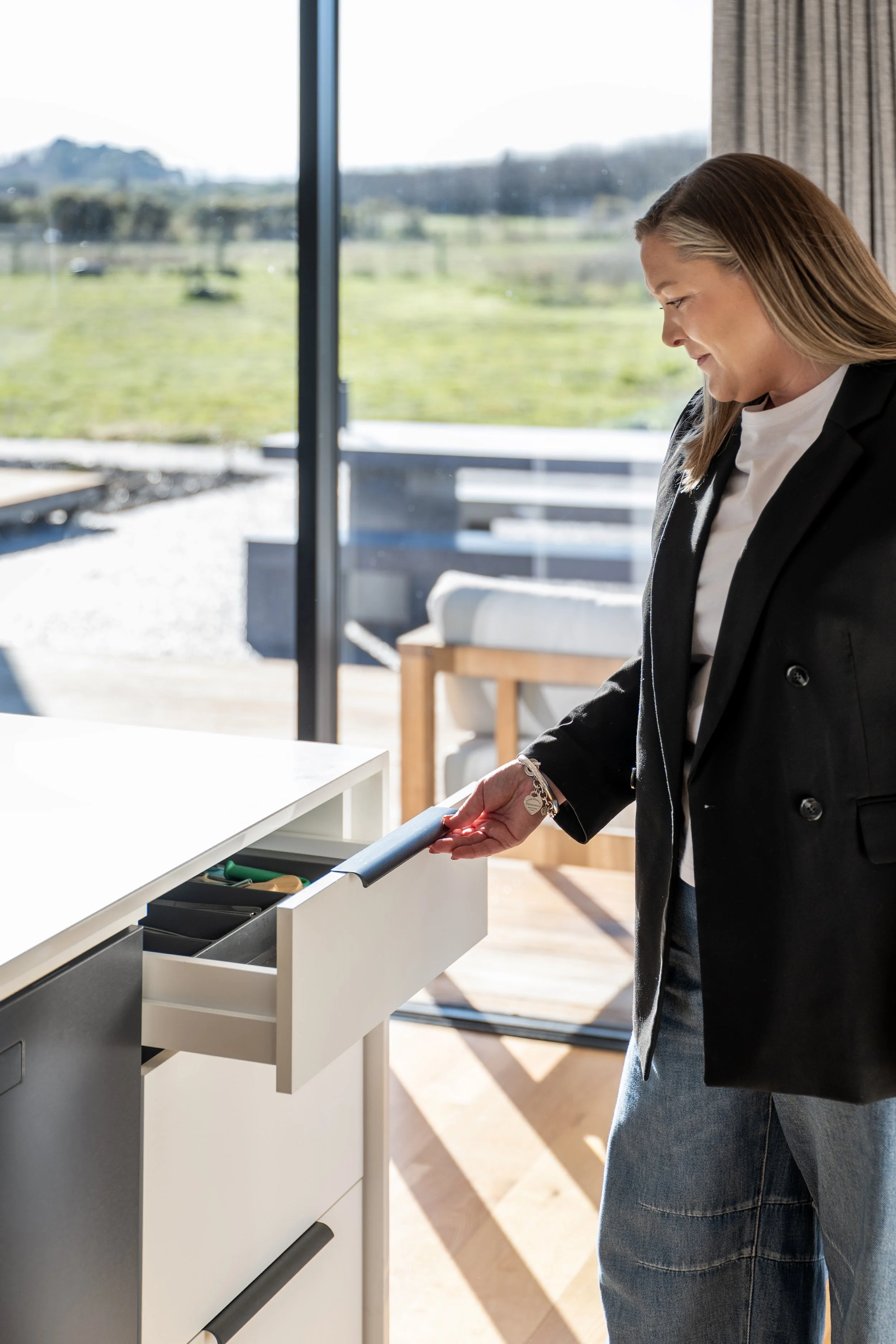 A woman in a black blazer and jeans is using a drawer in a white cabinet, with a view of a green outdoor landscape through large glass windows behind her. Renovations, Extensions & Additional Dwellings Canterbury | Alterhouse