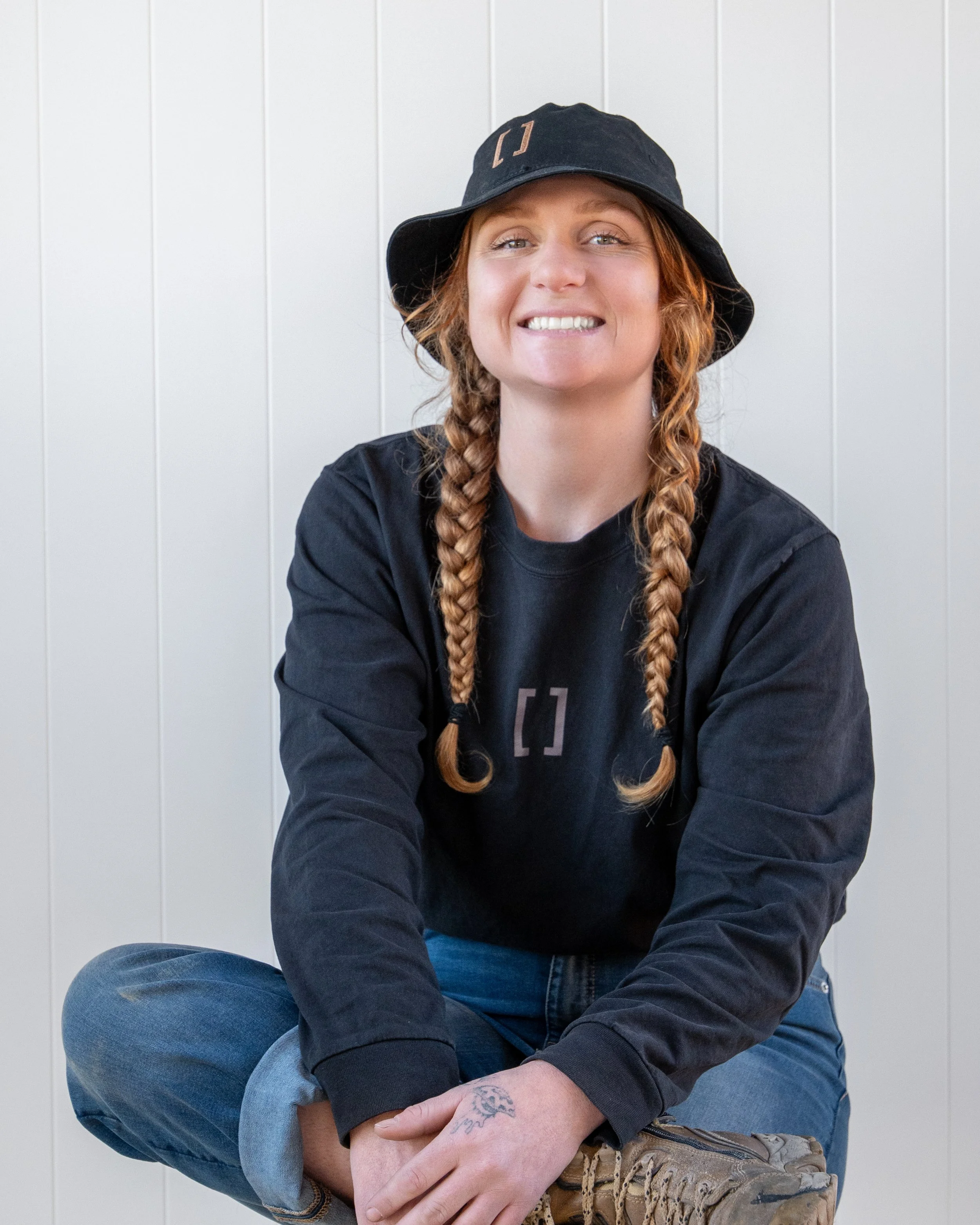 A woman with long braided red hair wearing a black bucket hat and black shirt, sitting against a white panel wall, smiling at the camera. Renovations, Extensions & Additional Dwellings Canterbury | Alterhouse