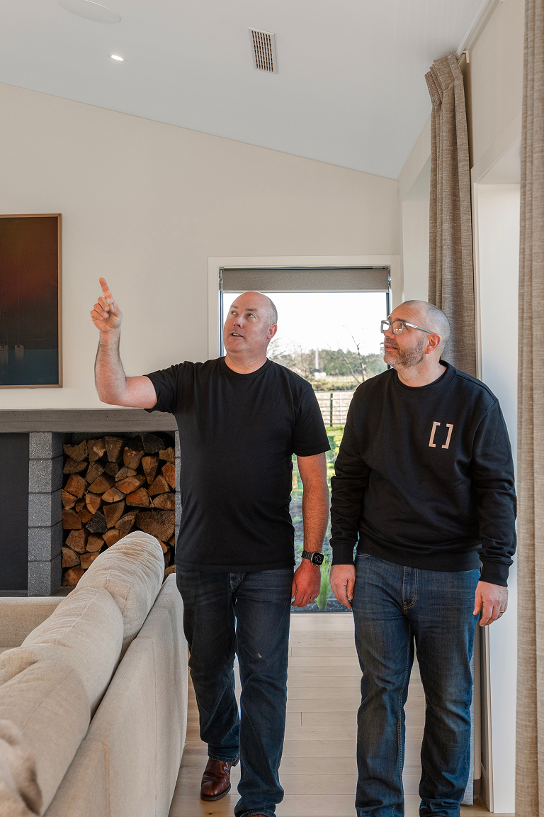 Two men walking inside a modern living room with a fireplace filled with stacked firewood, beige curtains, and a window showing an outdoor view. Renovations, Extensions & Additional Dwellings Canterbury | Alterhouse