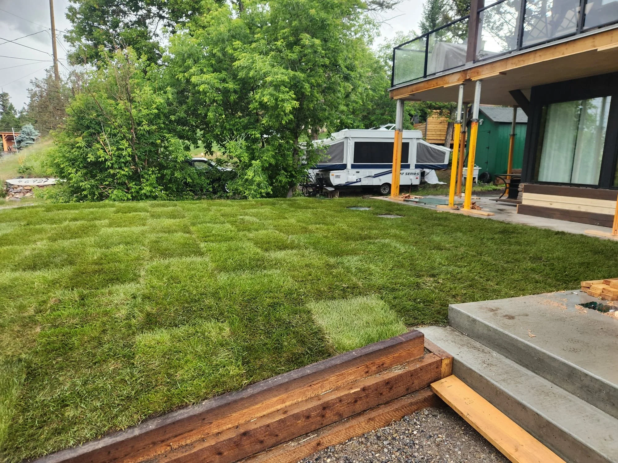 Residential backyard with green lawn, large leafy trees, a patio area under a house deck, and a white RV or camper parked behind the trees.