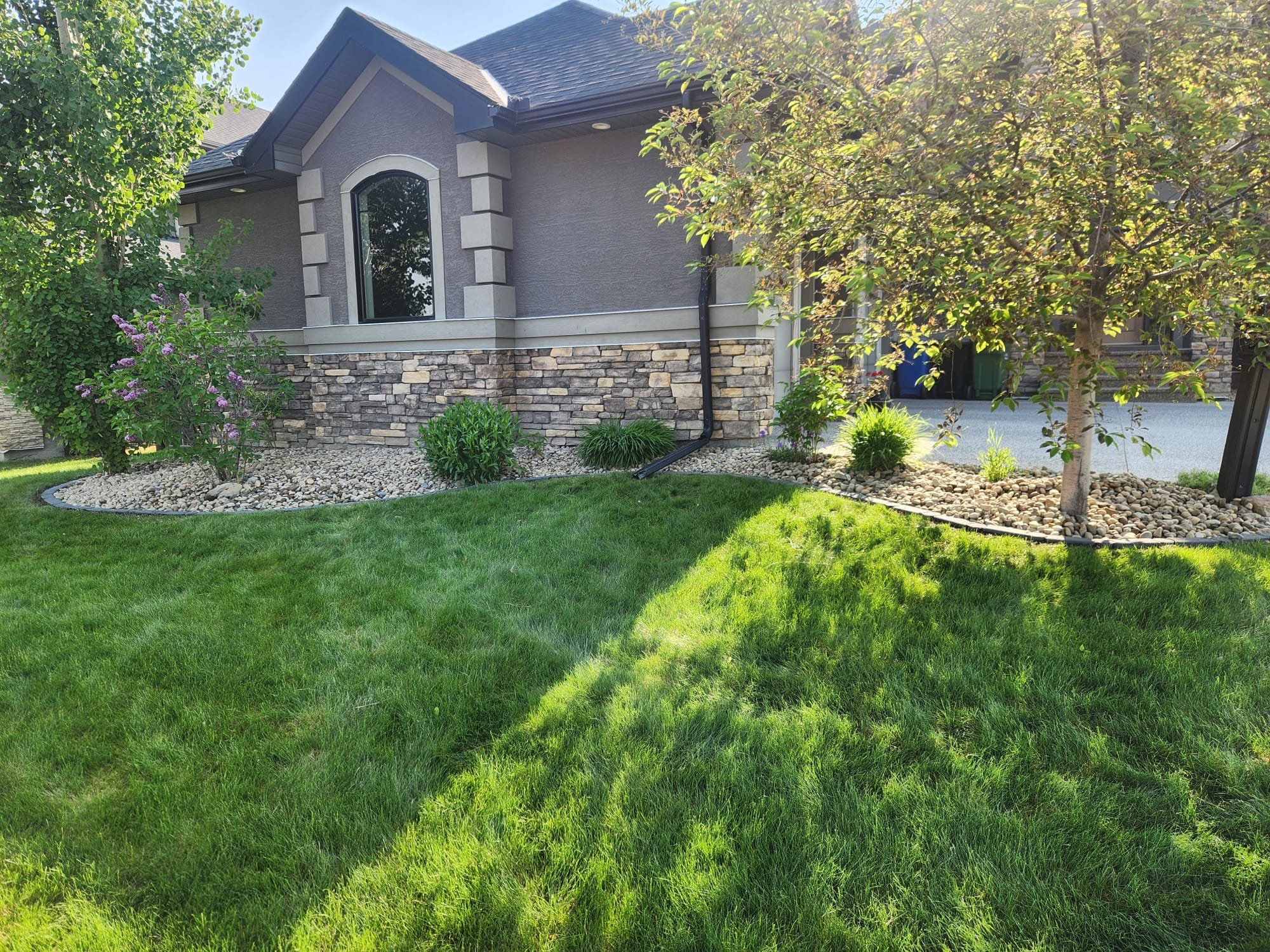 A well-maintained front yard with lush green grass, a small landscaped area with purple flowering bush, small green shrubs, and trees. A house with gray walls, stone accents at the bottom, arched window, and dark-colored roof is visible in the backgr
