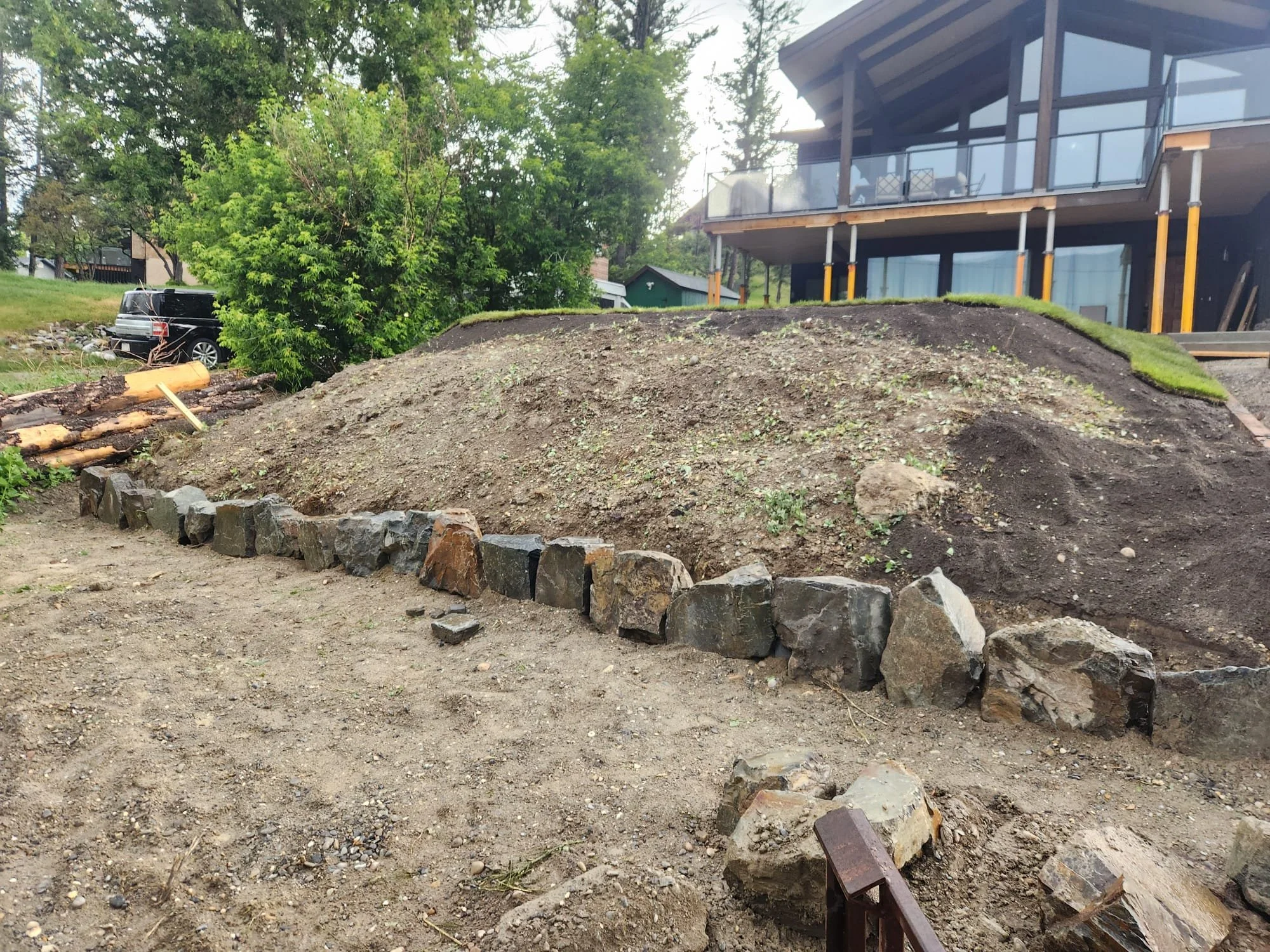 A backyard under construction with a dirt area bordered by a line of rocks, a small ramp leading up, and a modern house with large glass windows and a balcony in the background. There is a green tree on the left and a parked black vehicle behind it.