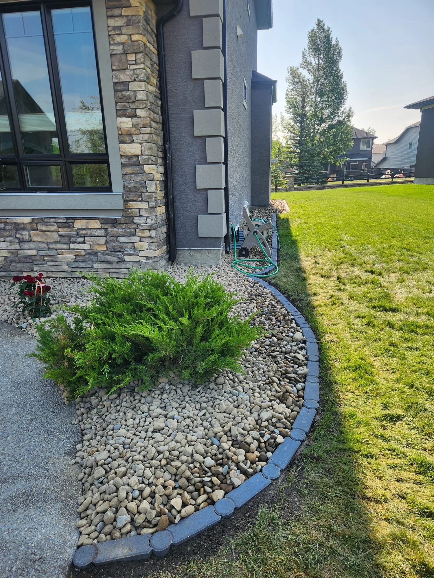 A landscaped yard with a house exterior on the left, featuring stone and gray siding, a large window, and a garden bed with small rocks, green shrubs, and a curved stone border. There's a hose reel and garden hose on the wall, and a large grassy lawn