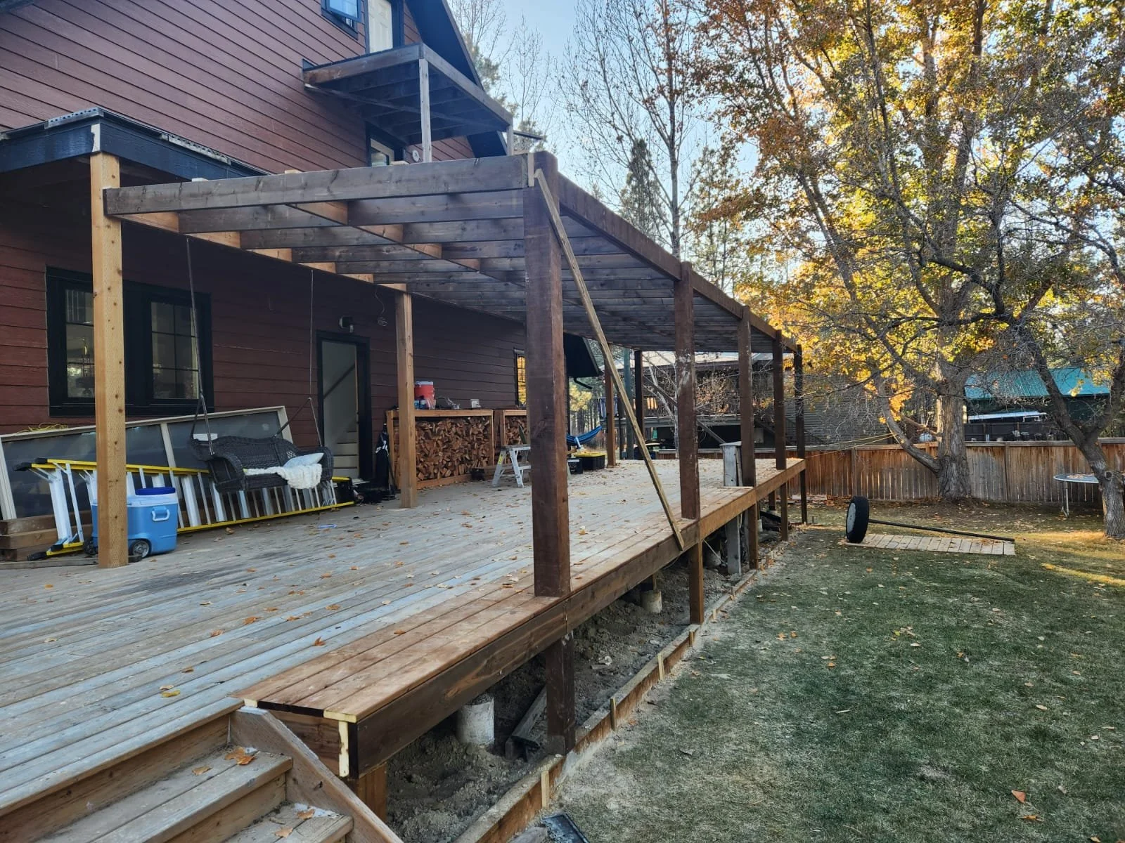 A construction site of a wooden deck attached to a house, with tools and materials stored on it, and a ladder leaning against the deck, in a backyard with trees and a fence.