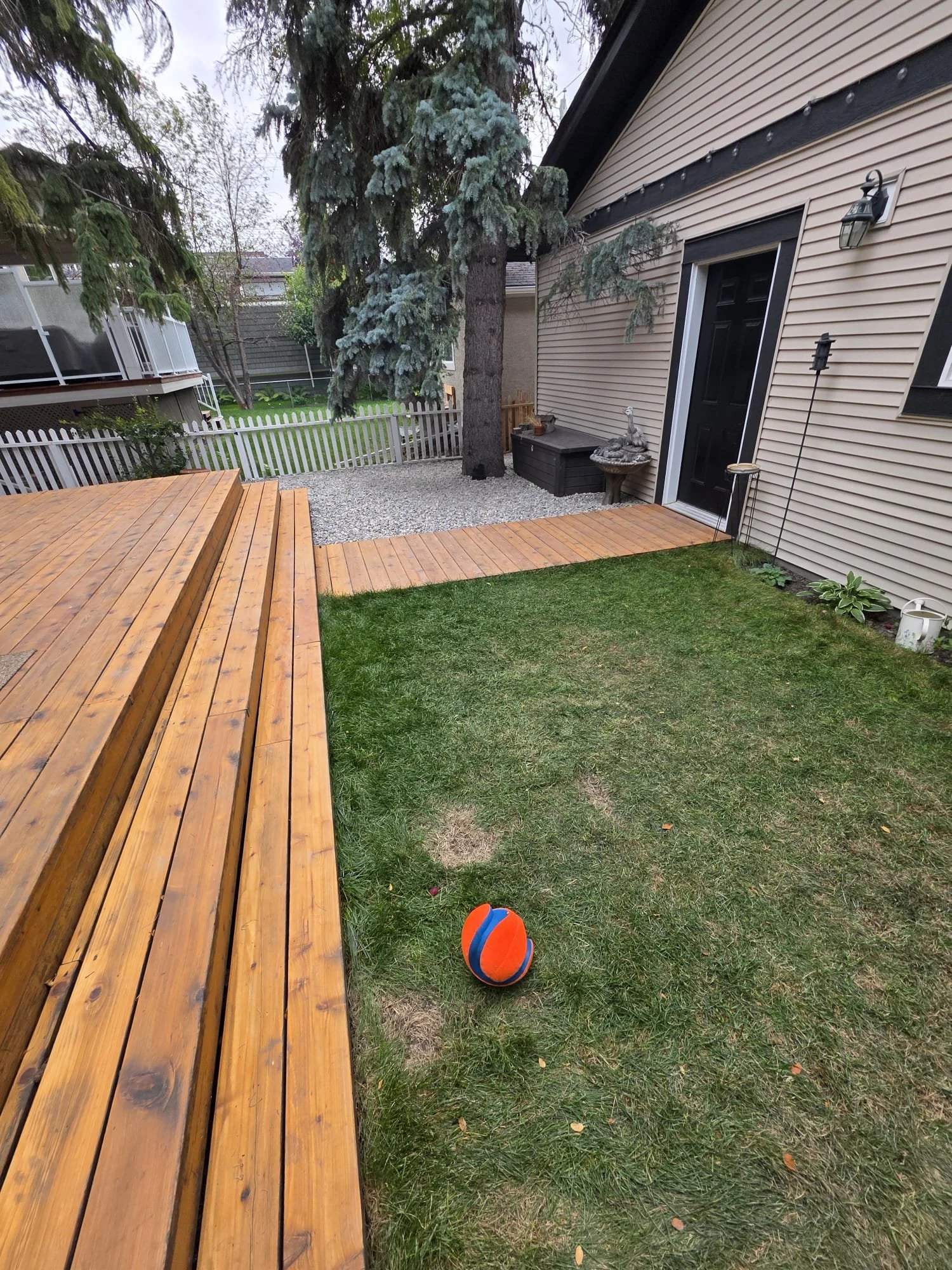 Backyard with a wooden deck, a grassy area, a large tree with drooping branches, a black door, a small statue on a pedestal, a fenced yard, and a ball on the grass.
