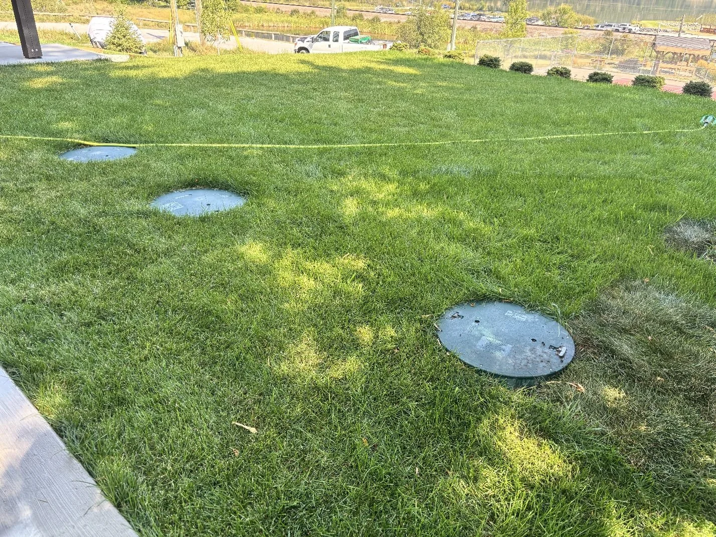 A grassy area with four underground utility access covers, some grass overgrown around them, under a sunny sky. In the background, there are vehicles and some small bushes.