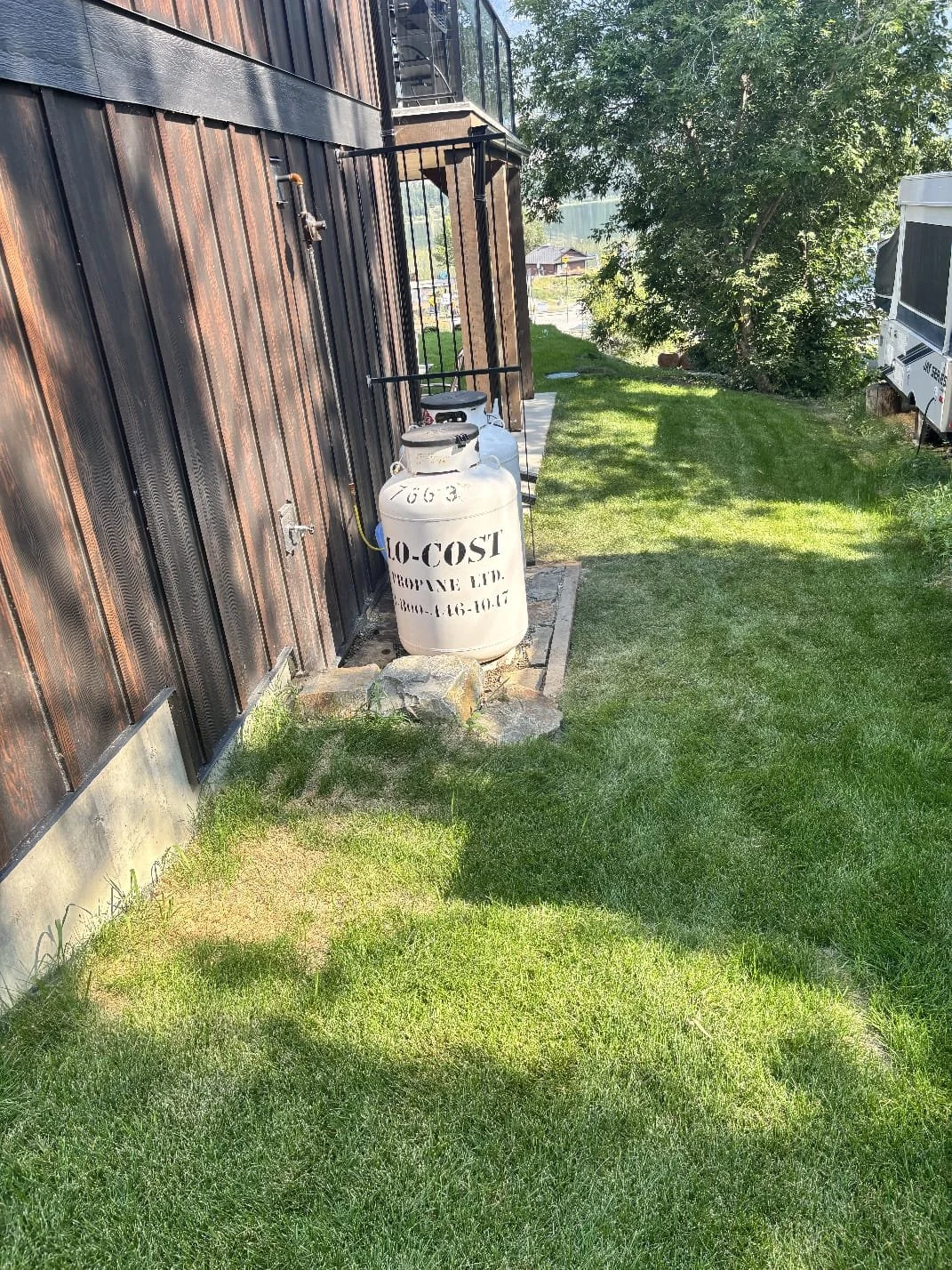 A grassy area next to a building with metal siding, featuring propane tanks labeled and a small stone patio, with trees and other buildings in the background.