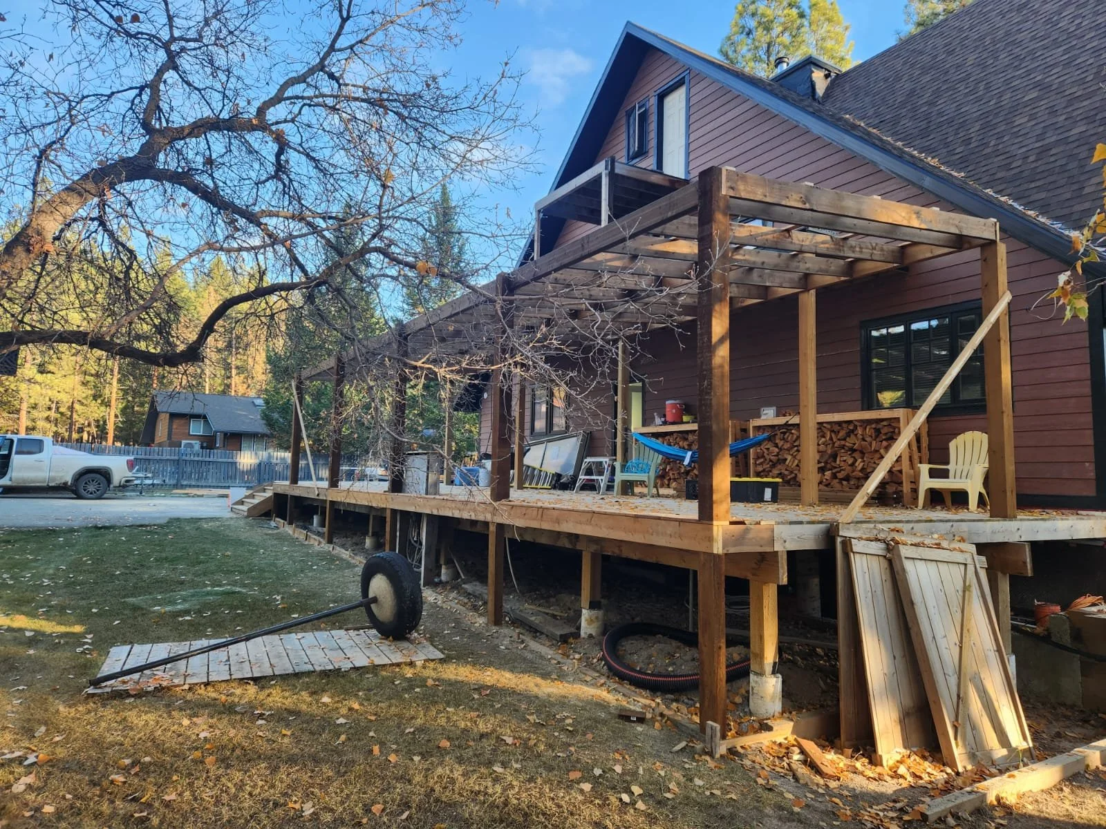 Bare wooden deck under construction attached to a brown house. The deck extends from the house with some furniture and firewood stacked on it. A section of the deck is missing, with construction materials and tools nearby, and a large tire attached t