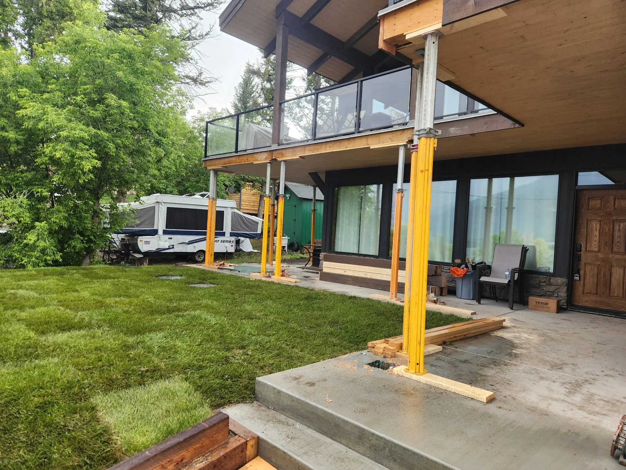 Exterior view of a house under construction with a new balcony and concrete steps, supported by yellow and silver metal scaffolding, in a lush green backyard with trees and a camping trailer.