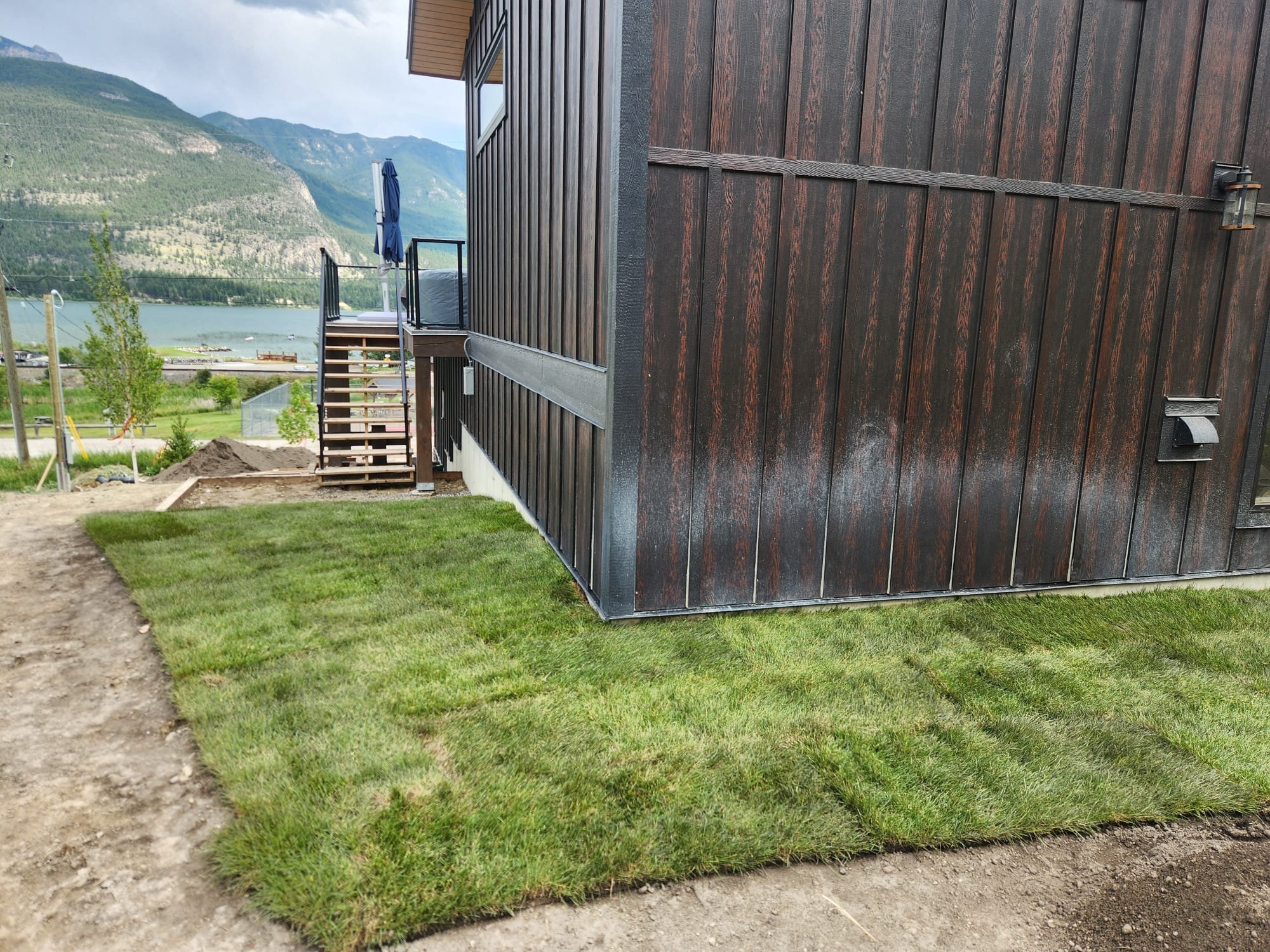 Side of a house with dark brown wooden vertical siding, a small staircase leading to a balcony with a blue umbrella, overlooking a lake and green mountainous landscape in the background.