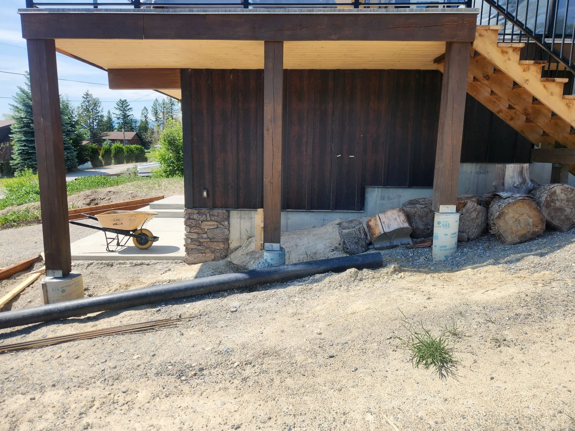A construction site with a building under construction. The building has black wooden siding with stone foundation and a wooden staircase. Construction materials like logs and buckets are present. The surrounding area has dirt and gravel, with trees 