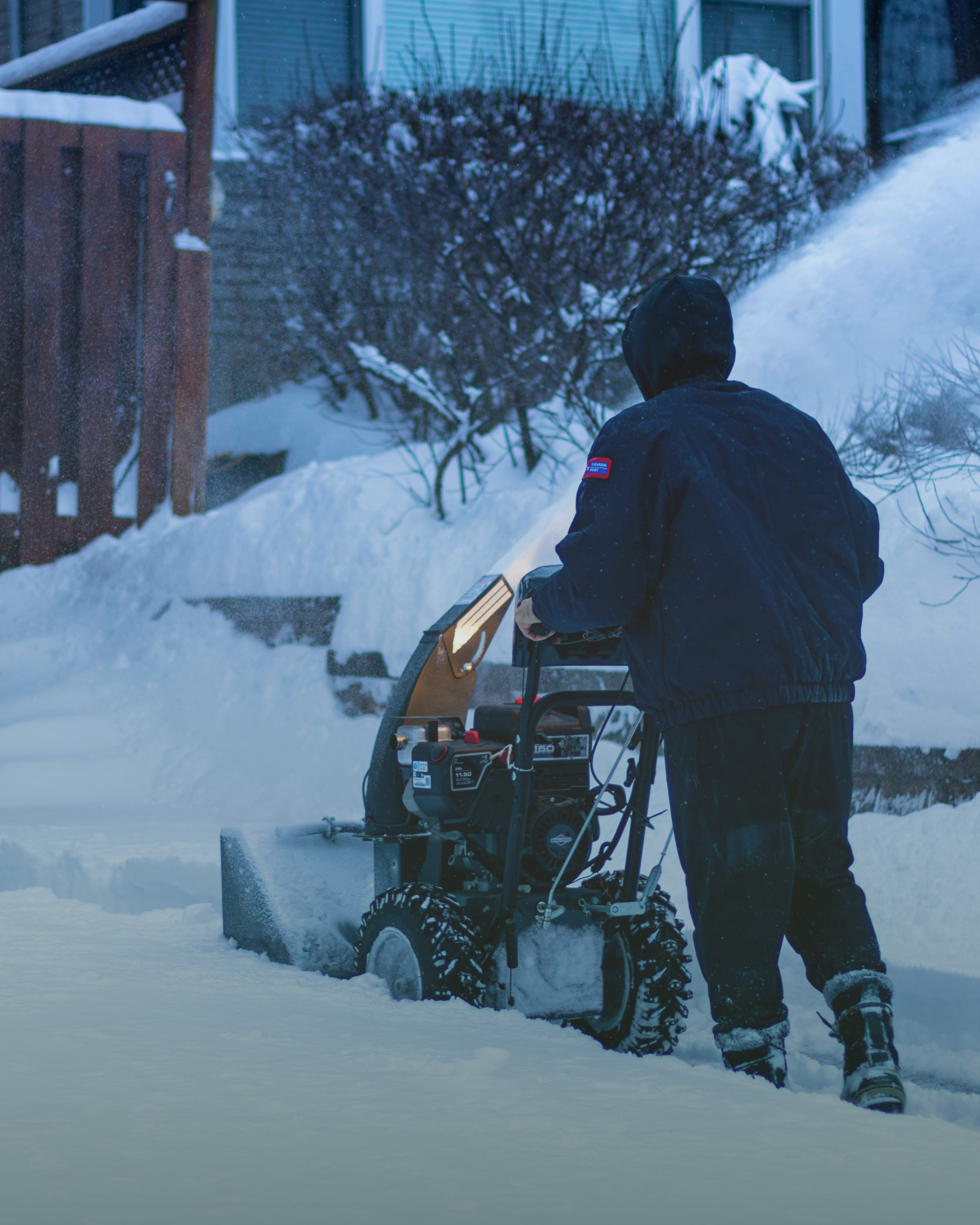 A person operating a snowblower in a residential yard during winter, with snow-covered bushes and a house in the background.