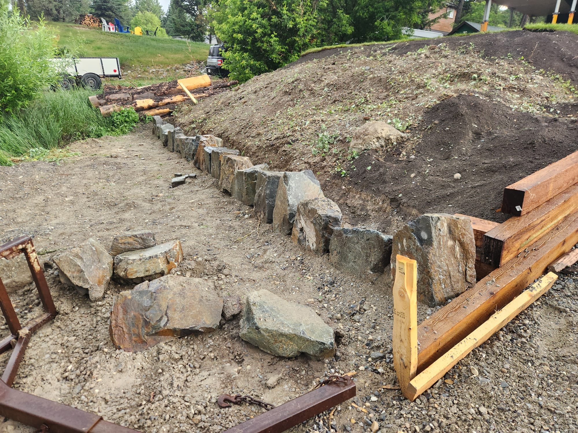 Construction site with a stone border along a dirt pathway. There are wooden beams, an earth embankment, piles of soil, and rocks, with trees and parked vehicles in the background.