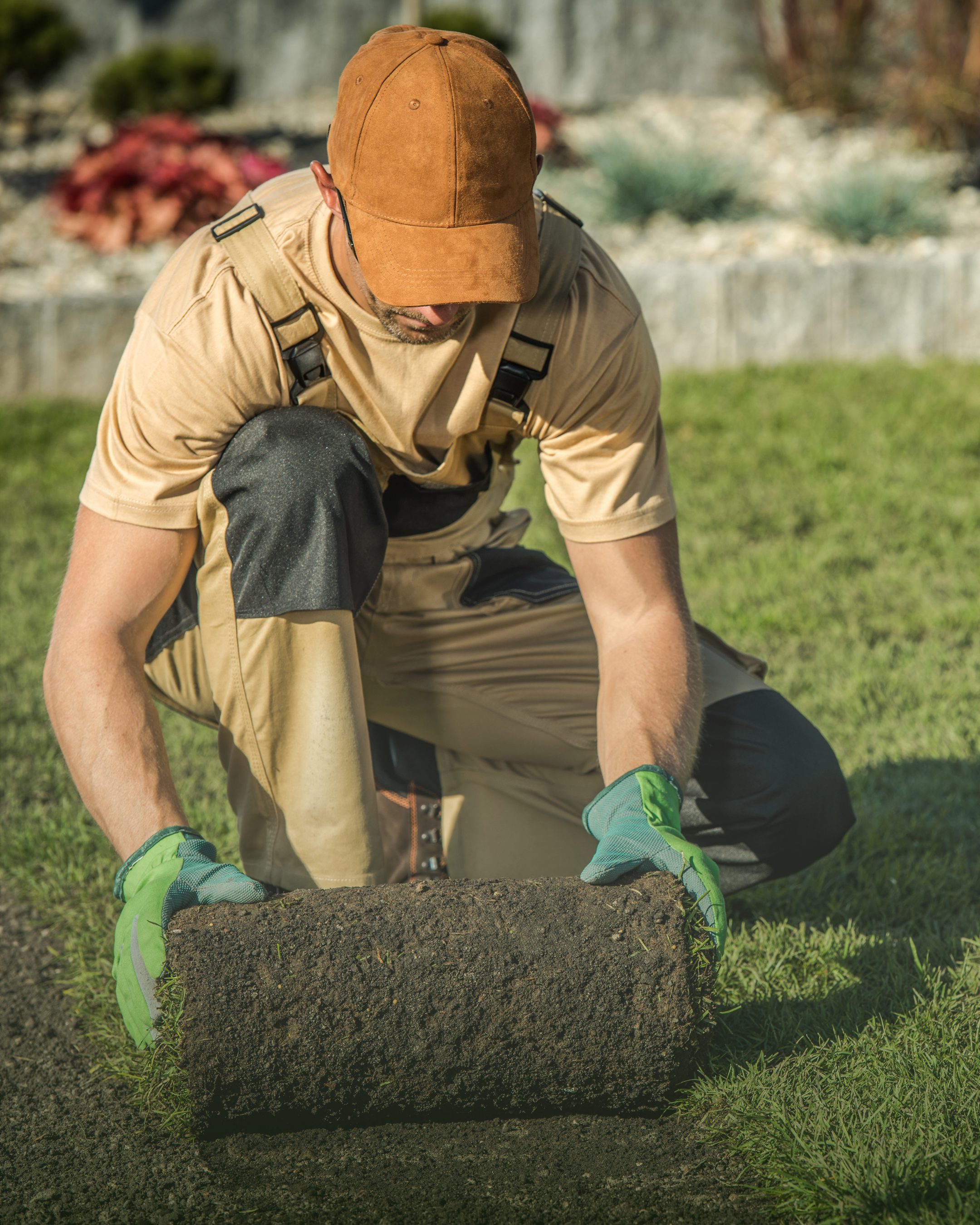 A man wearing a brown cap, tan shirt, and gloves is kneeling on the grass, pushing a large, freshly dug mound of soil.