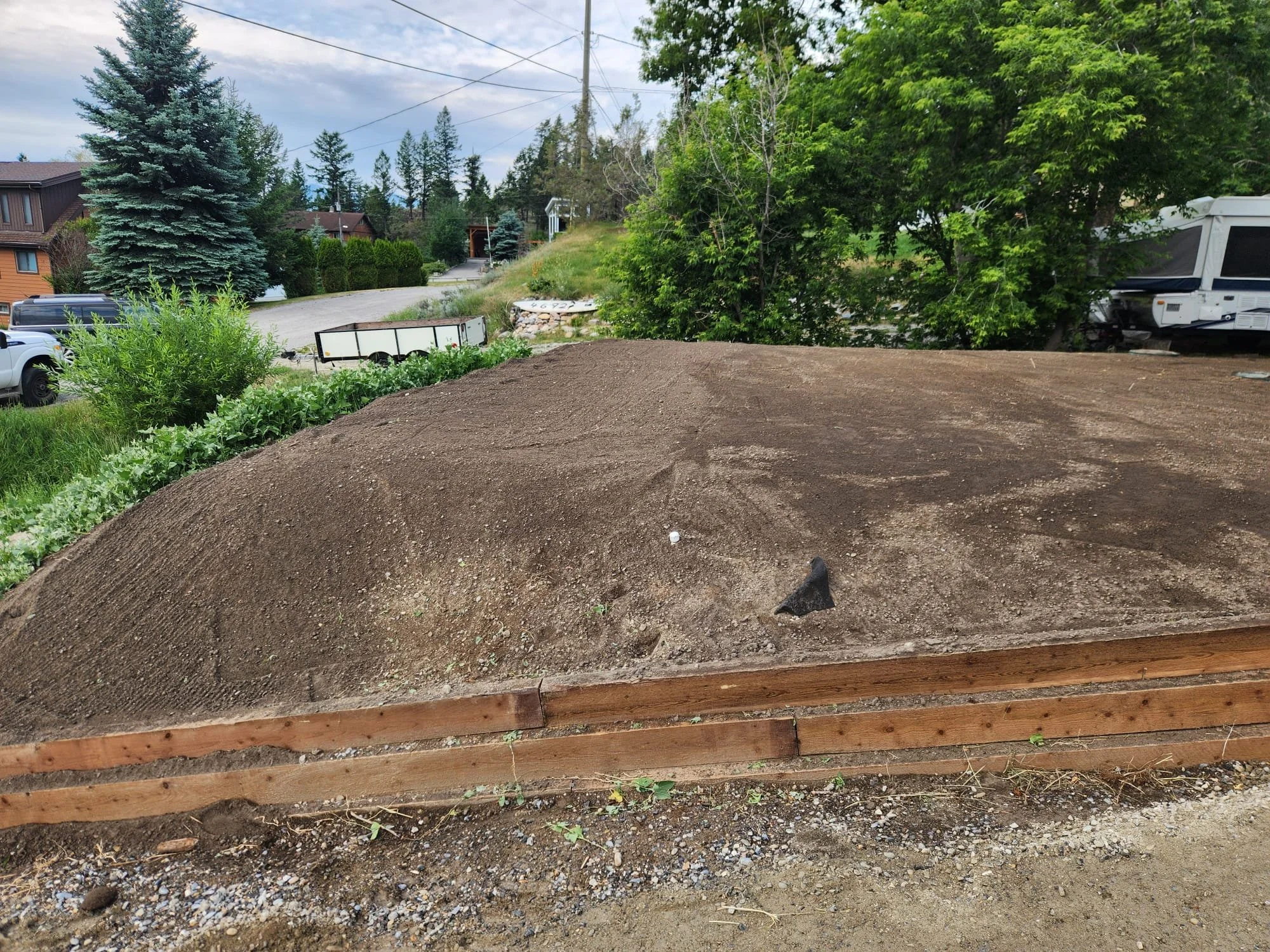 A backyard outdoor scene with freshly tilled soil in the foreground, green trees and bushes, and houses in the background. Part of a wooden frame surrounds the soil area, possibly for a garden bed.
