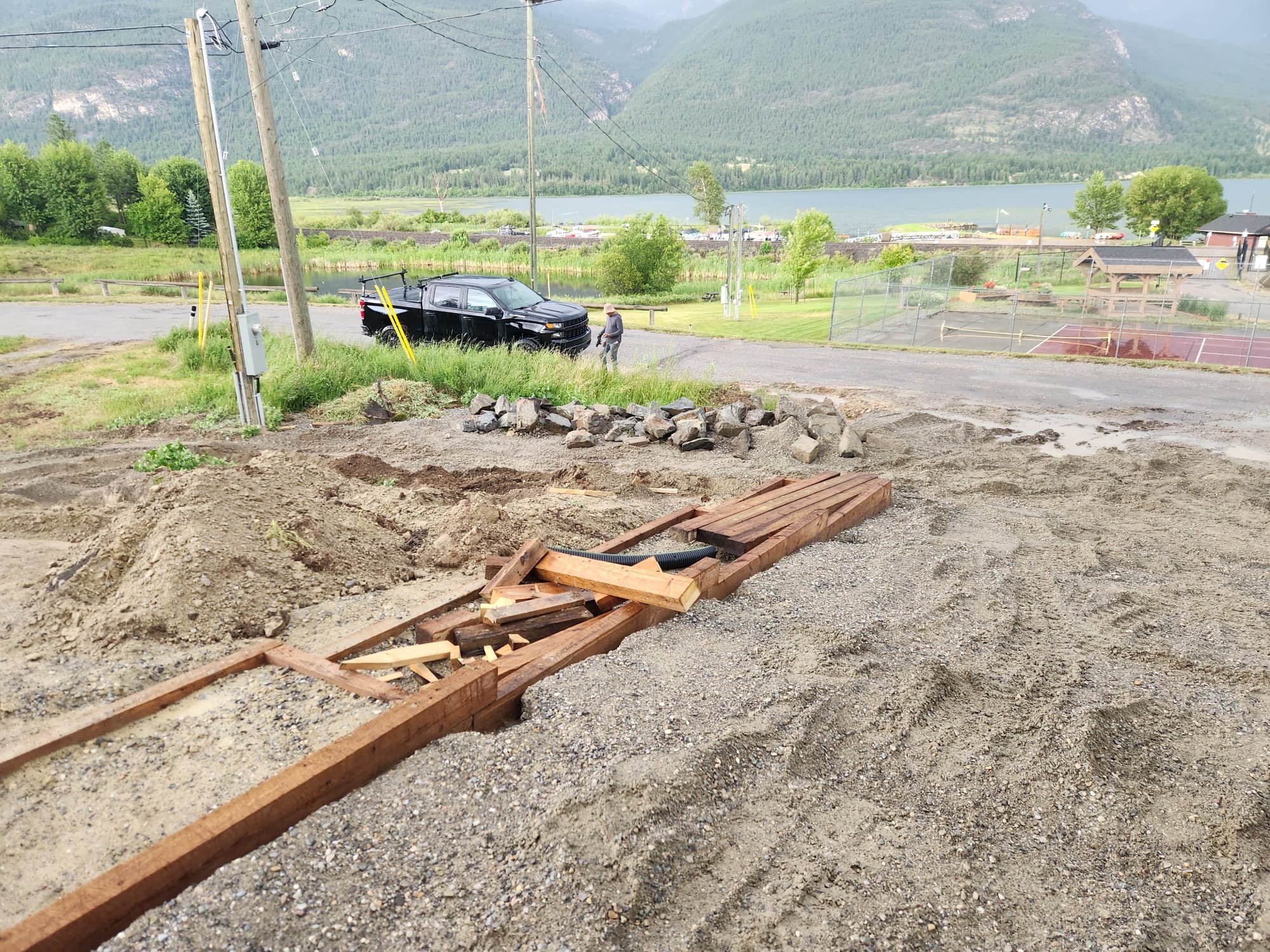 Construction site with dirt and gravel ground, wooden planks, and a partially constructed wooden frame in the foreground. In the background, there is a black pickup truck, a person walking, green trees, a tennis court, a lake, mountains, and a cloudy