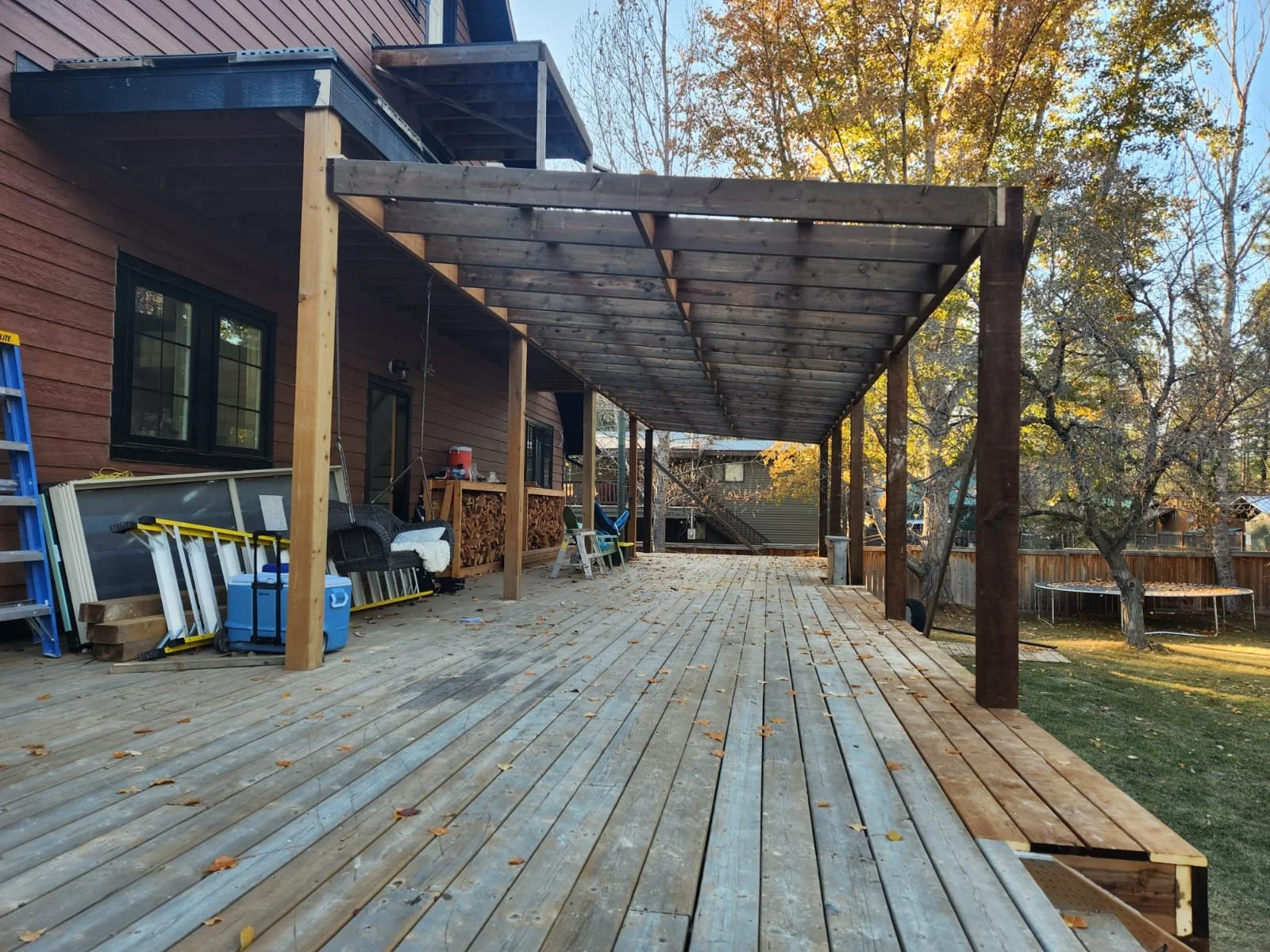 The image shows a backyard with a wooden deck under construction, featuring a partially built pergola with wooden beams and posts. There are tools, a ladder, and construction supplies on the deck. The house in the background has horizontal siding and