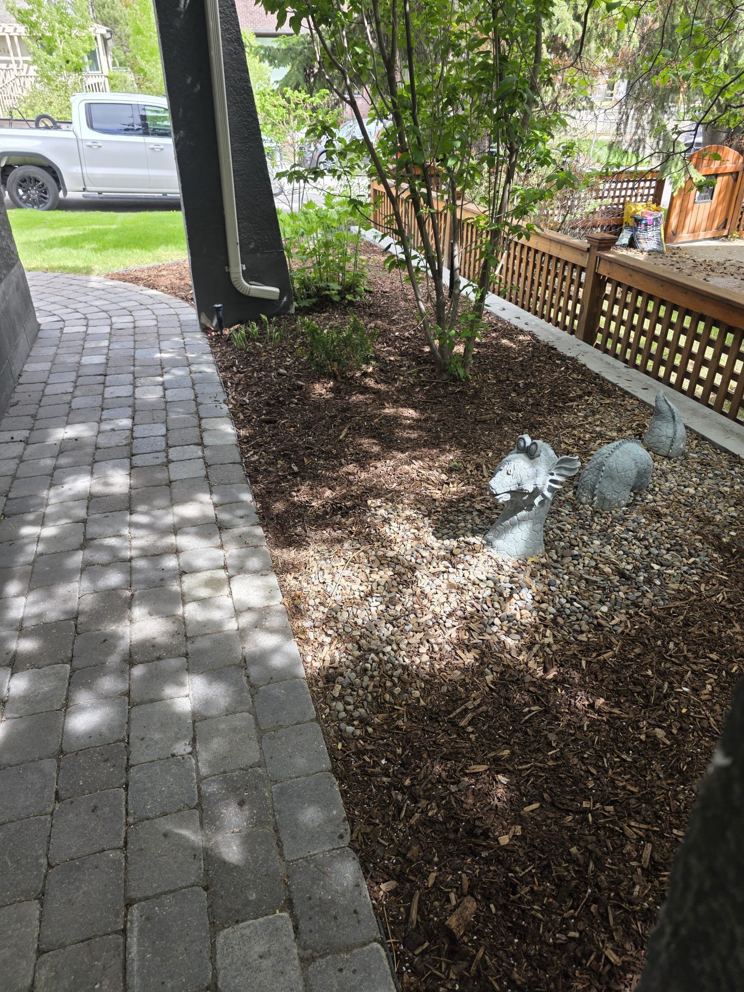 A garden area with a curved stone pathway, trees, bushes, and decorative animal garden ornaments. A wooden fence borders the garden, and a parked white pickup truck is visible in the background.