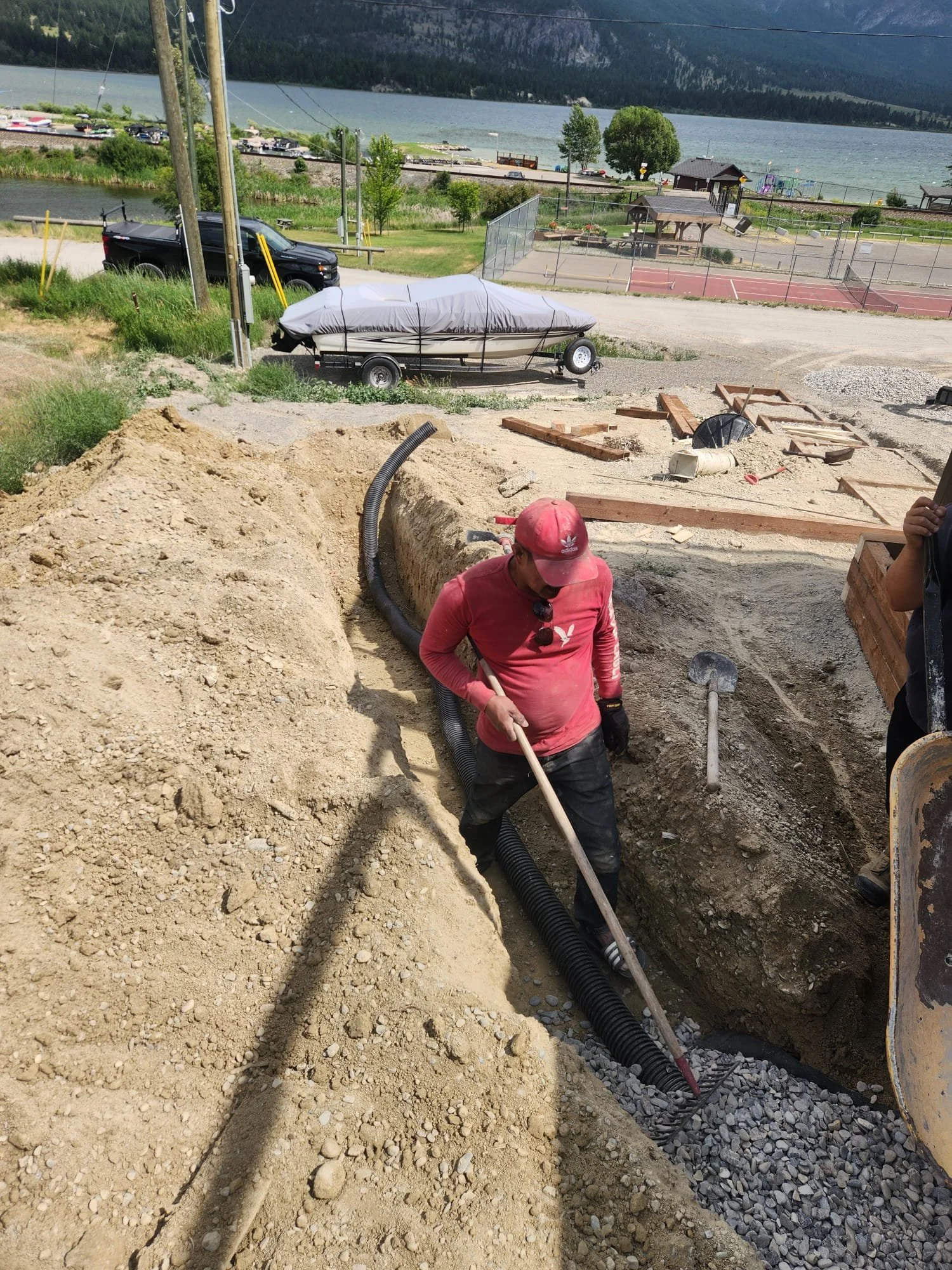 Construction workers dig a trench for underground pipes with a black corrugated pipe visible in foreground. A person in a red shirt and red cap is working in the trench, near construction materials and tools. In the background, a parked boat, a black