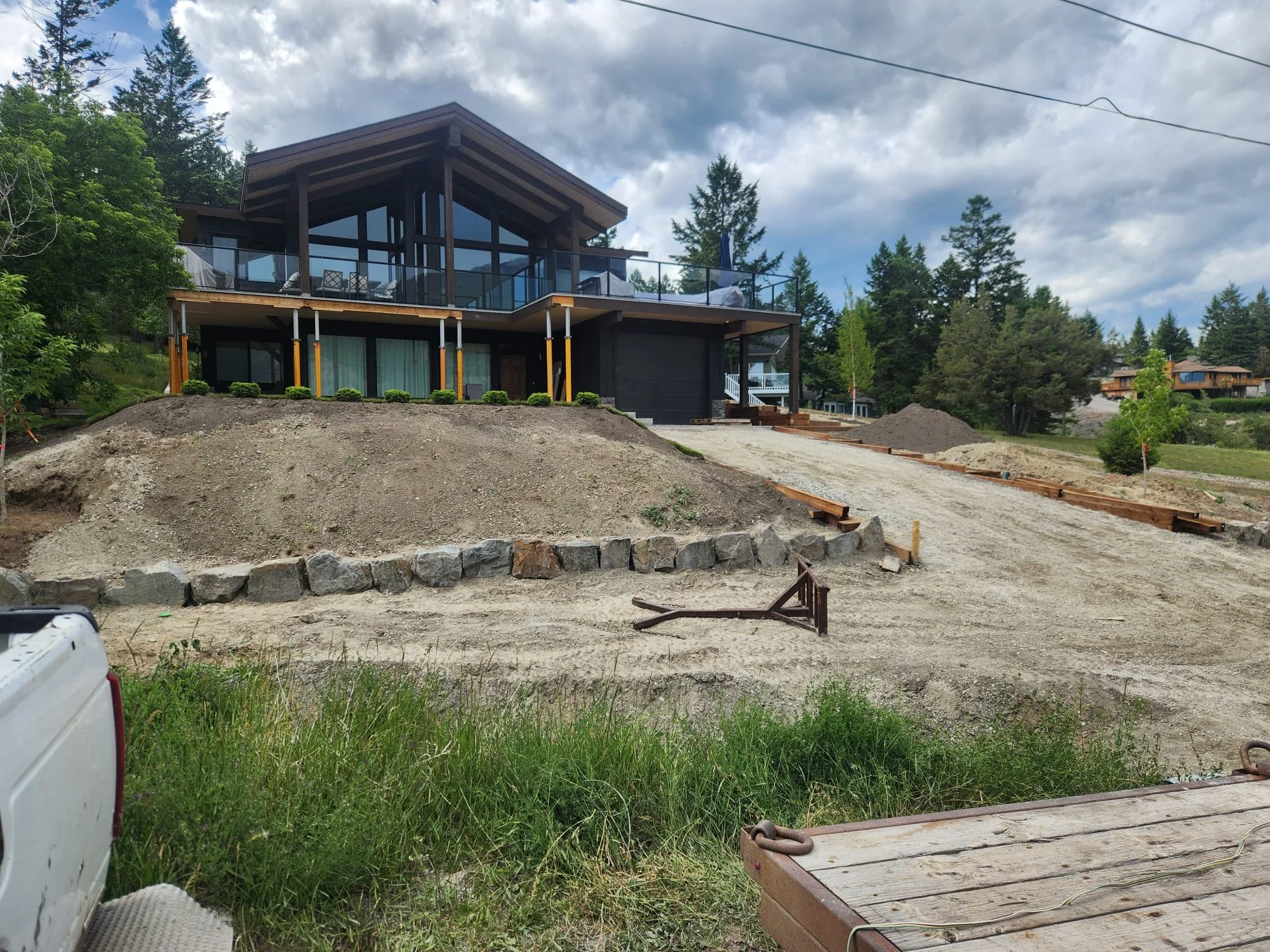 A modern two-story house with large glass windows and a balcony on the upper level, situated on a hill with a dirt driveway and landscaping in progress, surrounded by trees and a cloudy sky.