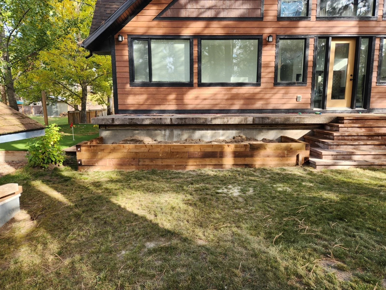 Backyard of a house with a new wooden deck under construction, featuring stairs leading up to a sliding glass door and large front windows. The area is surrounded by green trees and grass.
