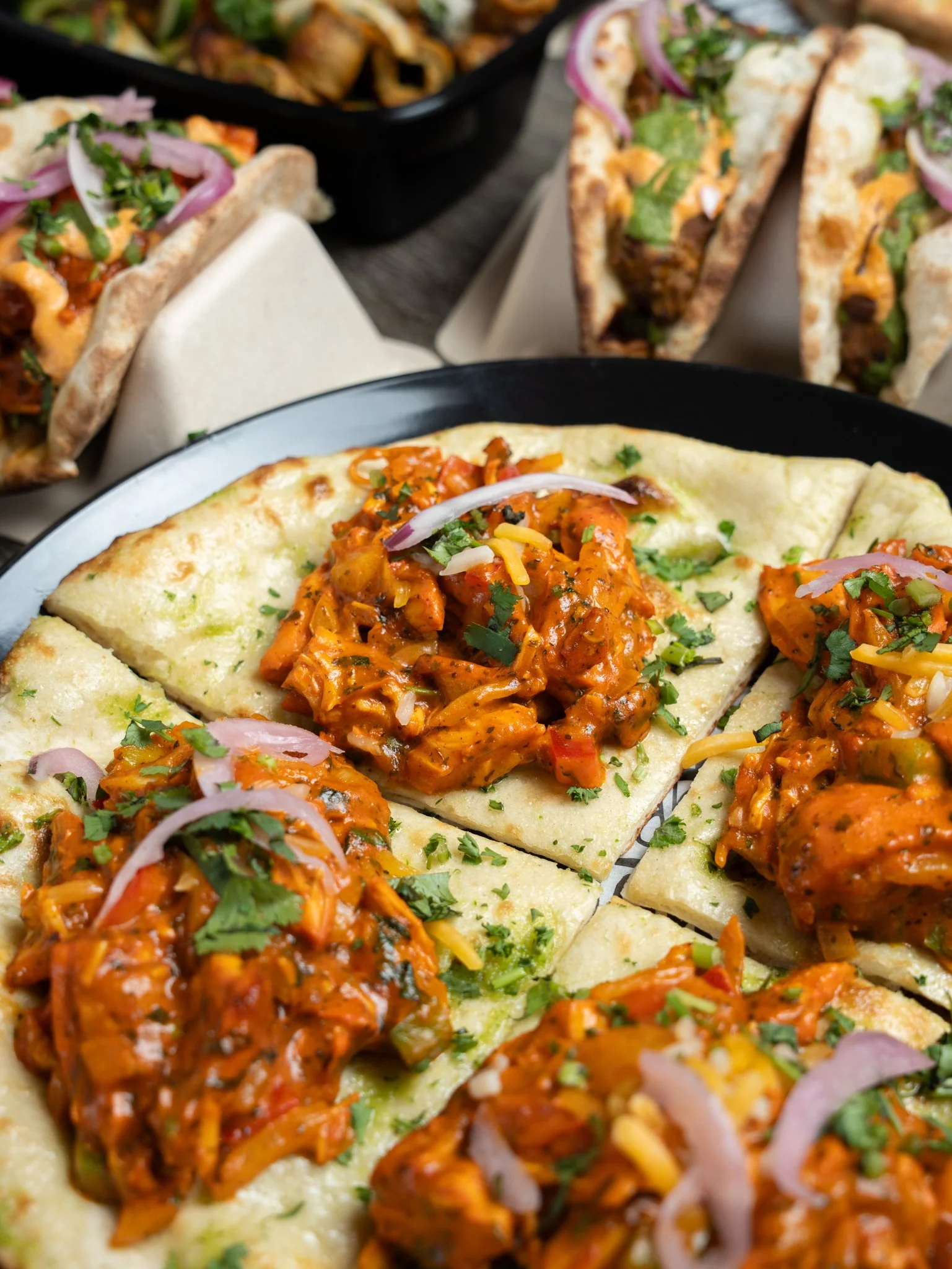 Close-up of a pizza topped with chicken, sauce, chopped herbs, and chopped onions, with additional slices of pizza and a tray of other foods in the background.