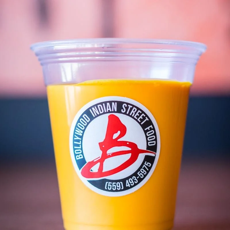 Cup of yellow mango lassi with a logo label from Bollywood Indian Street Food restaurant on the front.