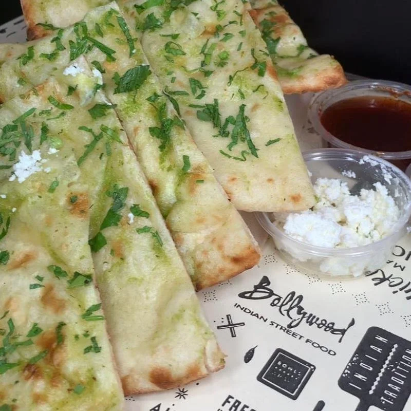 Four slices of cheese garlic naan bread garnished with chopped cilantro, served with small containers of brown dipping sauce and white ricotta cheese.