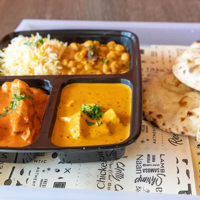 A meal with three compartments: rice with toppings, chickpeas or lentils curry, and butter chicken curry, served with naan bread on the side.