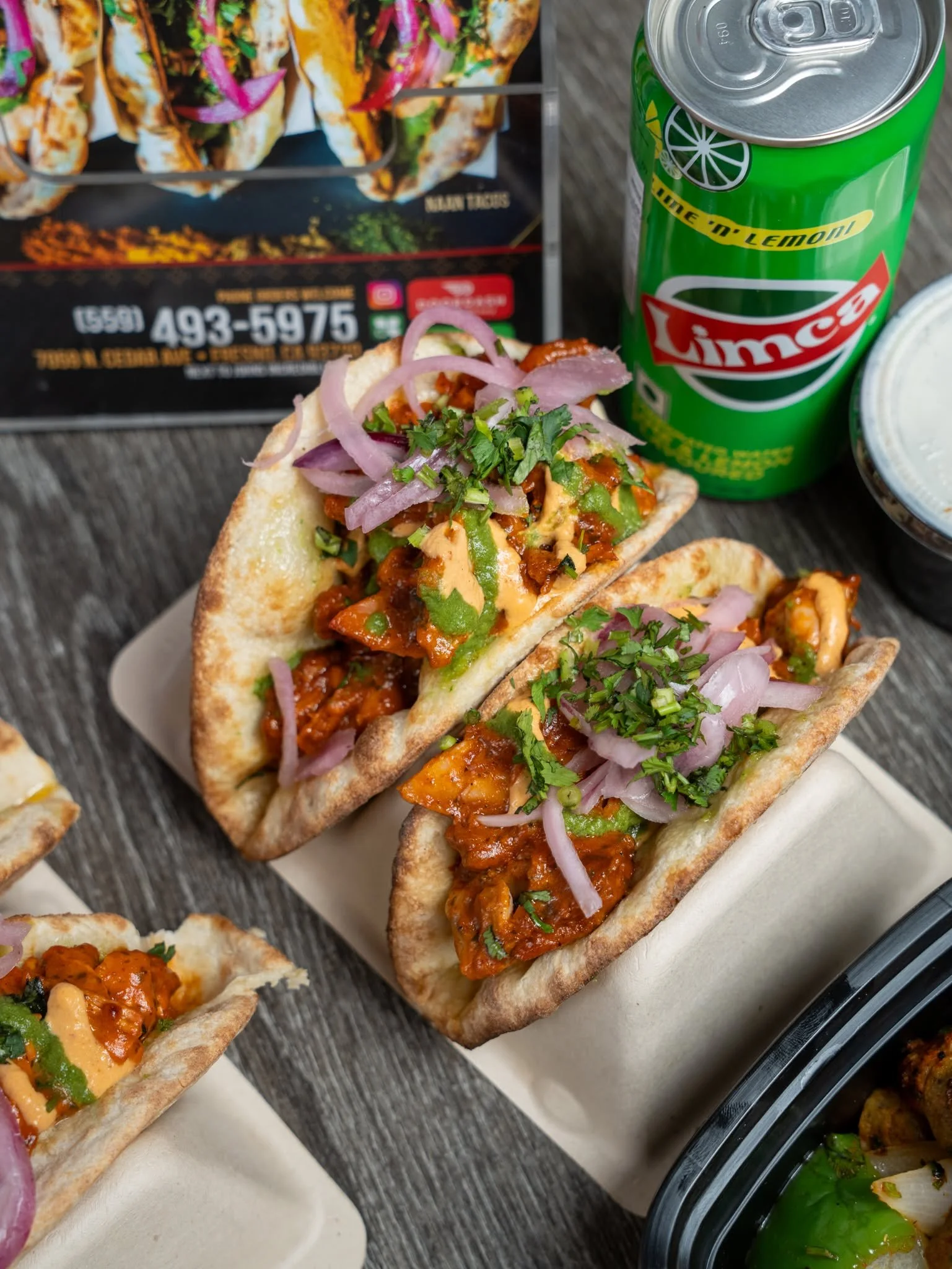 Small tacos filled with meat, topped with chopped onions and cilantro, placed on a white paper sheet on a dark wooden surface, with a green canned soda and other food items in the background.