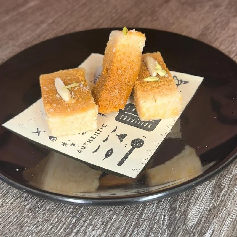Three small rectangular pieces of fried food garnished with slivered almonds on black plate lined with paper featuring black illustrations and text, on a wooden table.