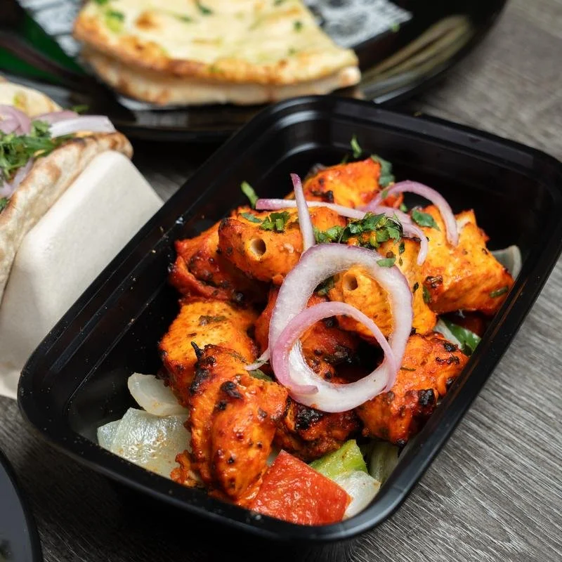 A black container with pieces of grilled or tandoori chicken garnished with sliced onions and cilantro, over chopped vegetables like onions and red peppers.