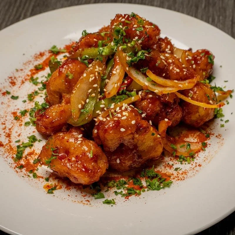 Plate of crispy chicken bites coated in sauce, garnished with chopped green onions, sesame seeds, and a sprinkle of red spice powder.
