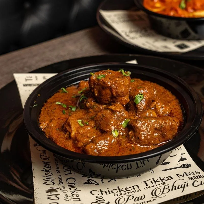 A black bowl filled with a serving of Indian chicken curry garnished with chopped cilantro, placed on a black tray with printed paper underneath, and another similar bowl of curry partially visible in the background.