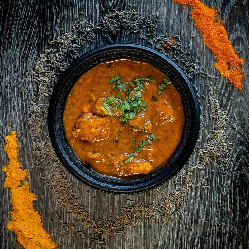A bowl of Indian curry garnished with cilantro, surrounded by cumin seeds and turmeric powder on a wooden surface.
