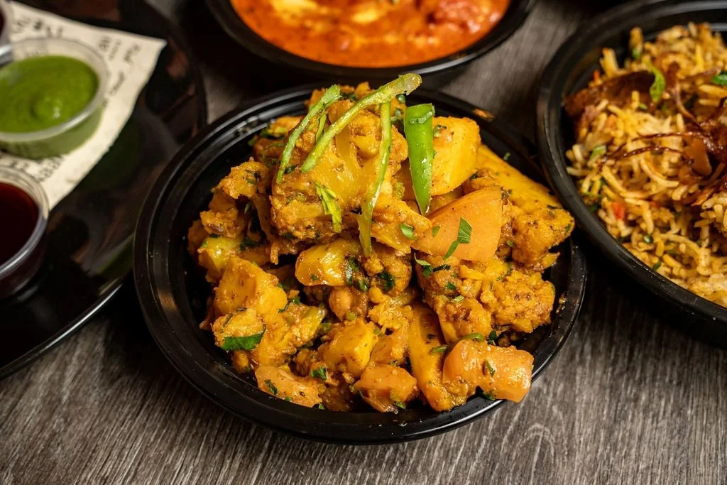 Close-up of a bowl of Indian chicken curry with mango chunks and green garnish, surrounded by other dishes including rice and sauces.