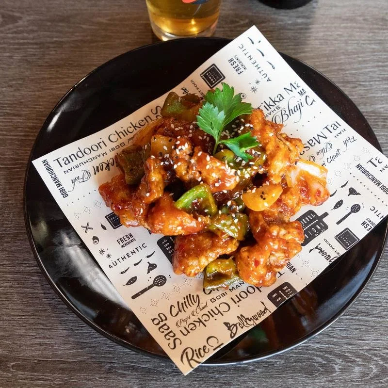 A black plate with sweet and sour chicken topped with sesame seeds and a sprig of cilantro, served on decorative paper, with a glass of beer in the background.