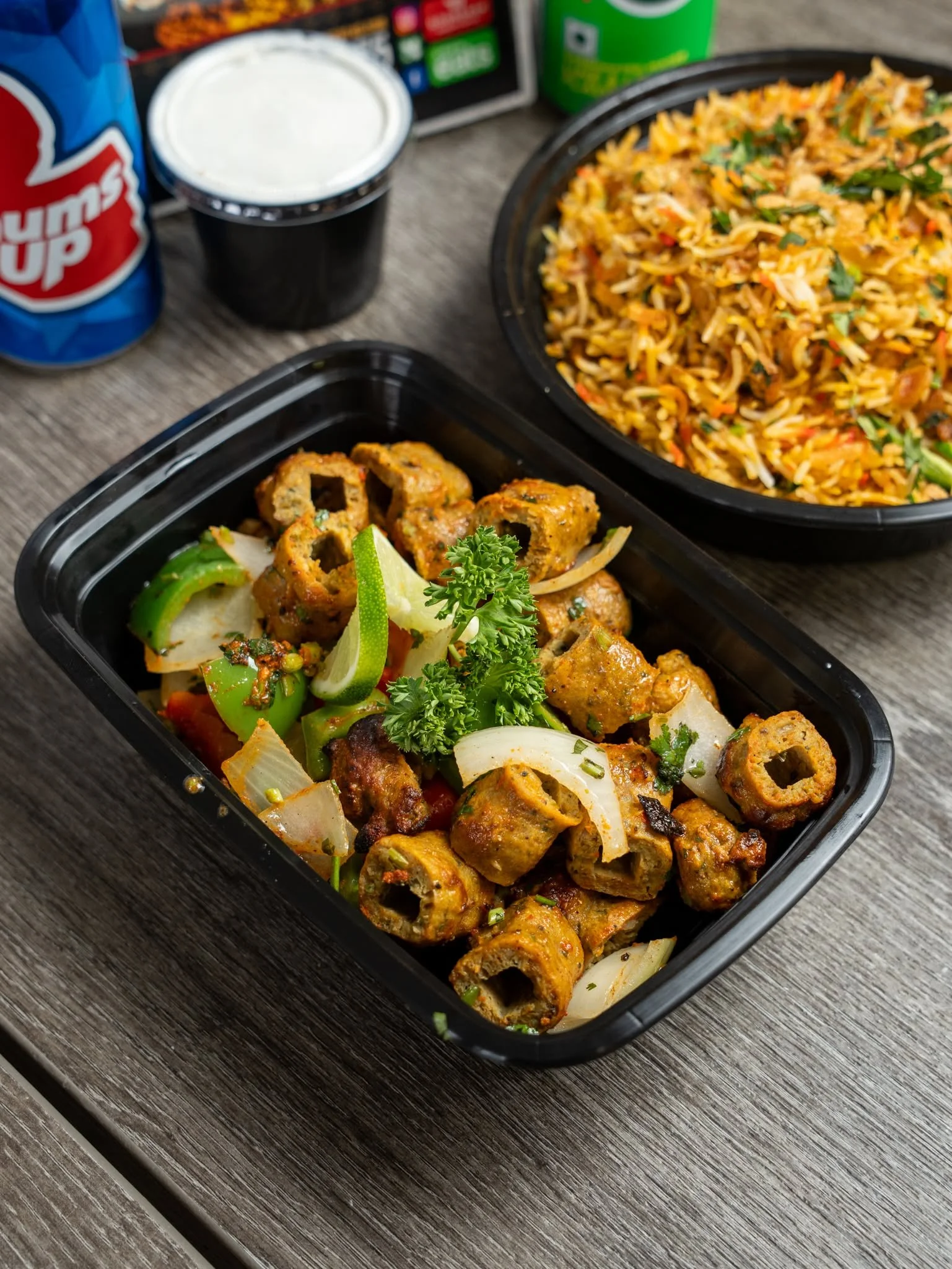 Takeout containers of Indian food with naan, rice, and meat, along with a soda and a container of yogurt, on a wooden table.