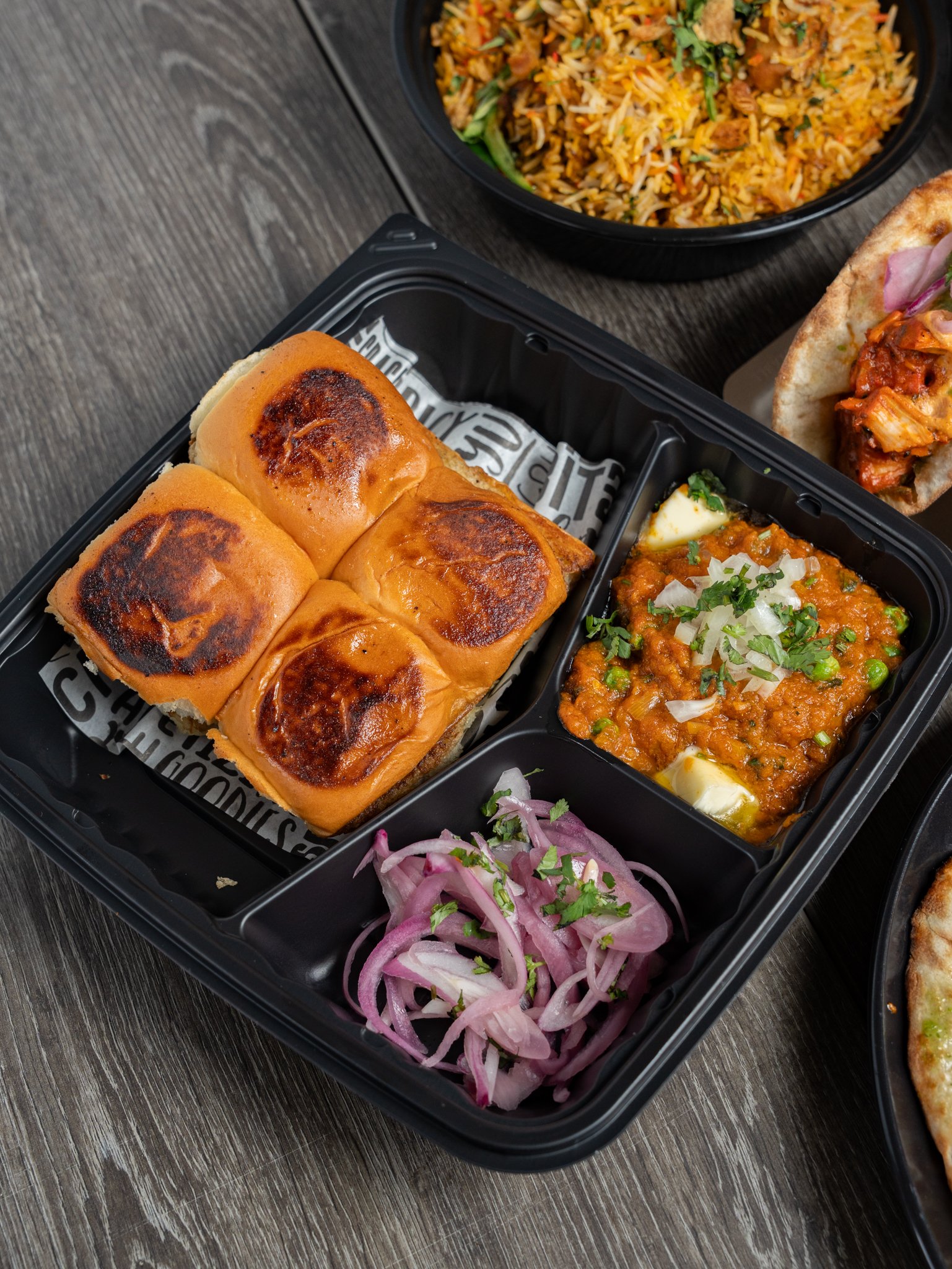 Takeout container with three bread rolls with browned tops, bowl of rice with vegetables and meat, side of sliced onion and cilantro, and a portion of curry with peas, carrots, and herbs.