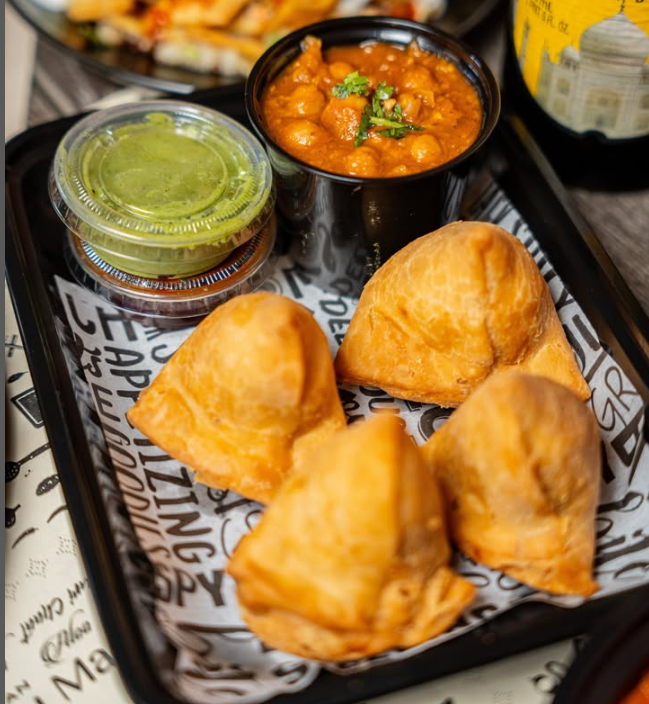 Four fried samosas served with green chutney and spicy chickpea curry with cilantro on a black tray.