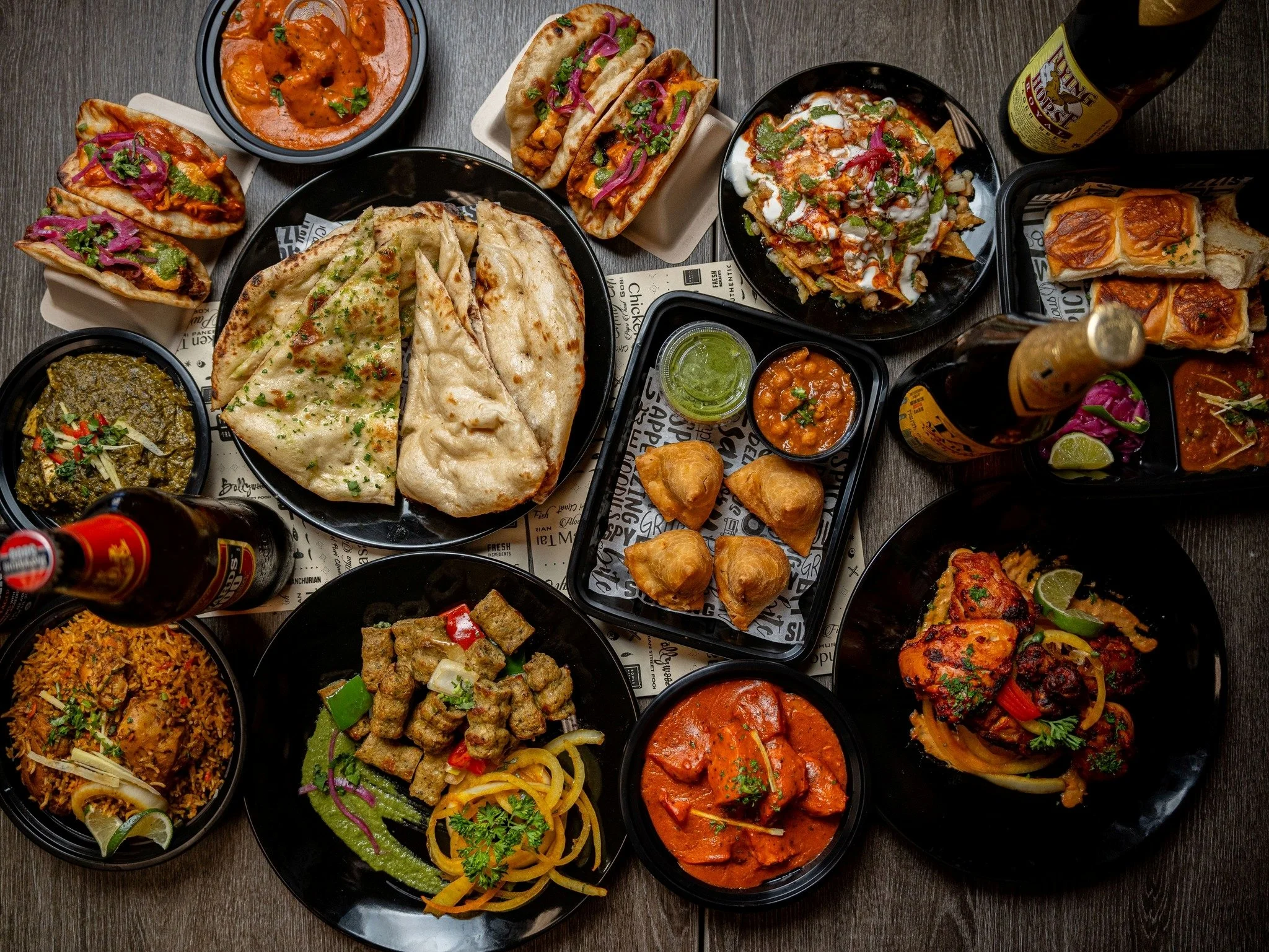 An array of Indian dishes including naan bread, rice, curry, tacos, nachos, and samosas, along with bottles of beer and small containers of sauces on a dark wooden table.