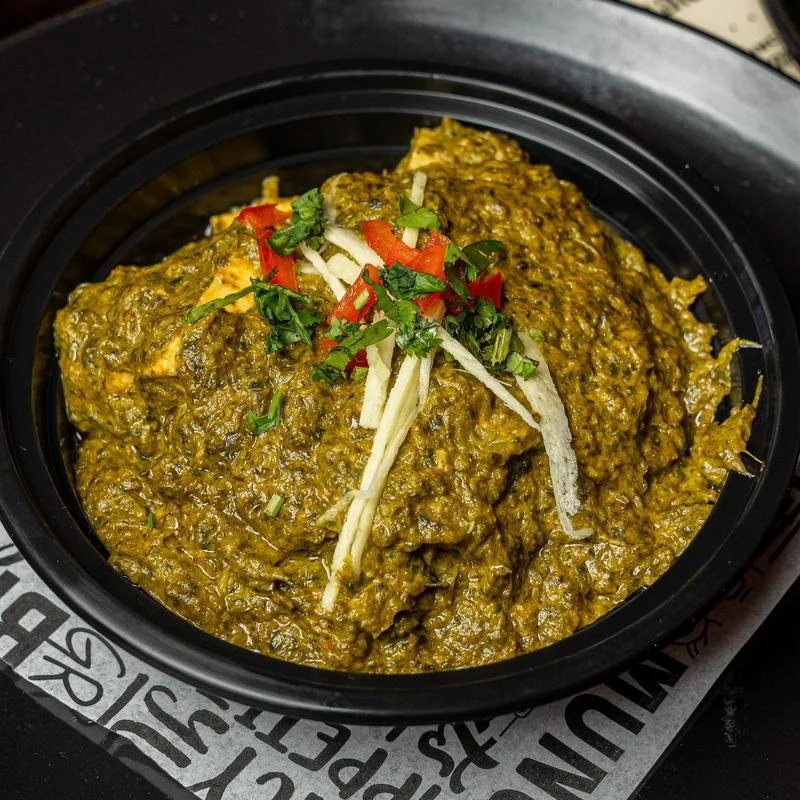 A black bowl of Indian spinach and meat curry garnished with chopped cilantro, shredded ginger, and red chili slices.