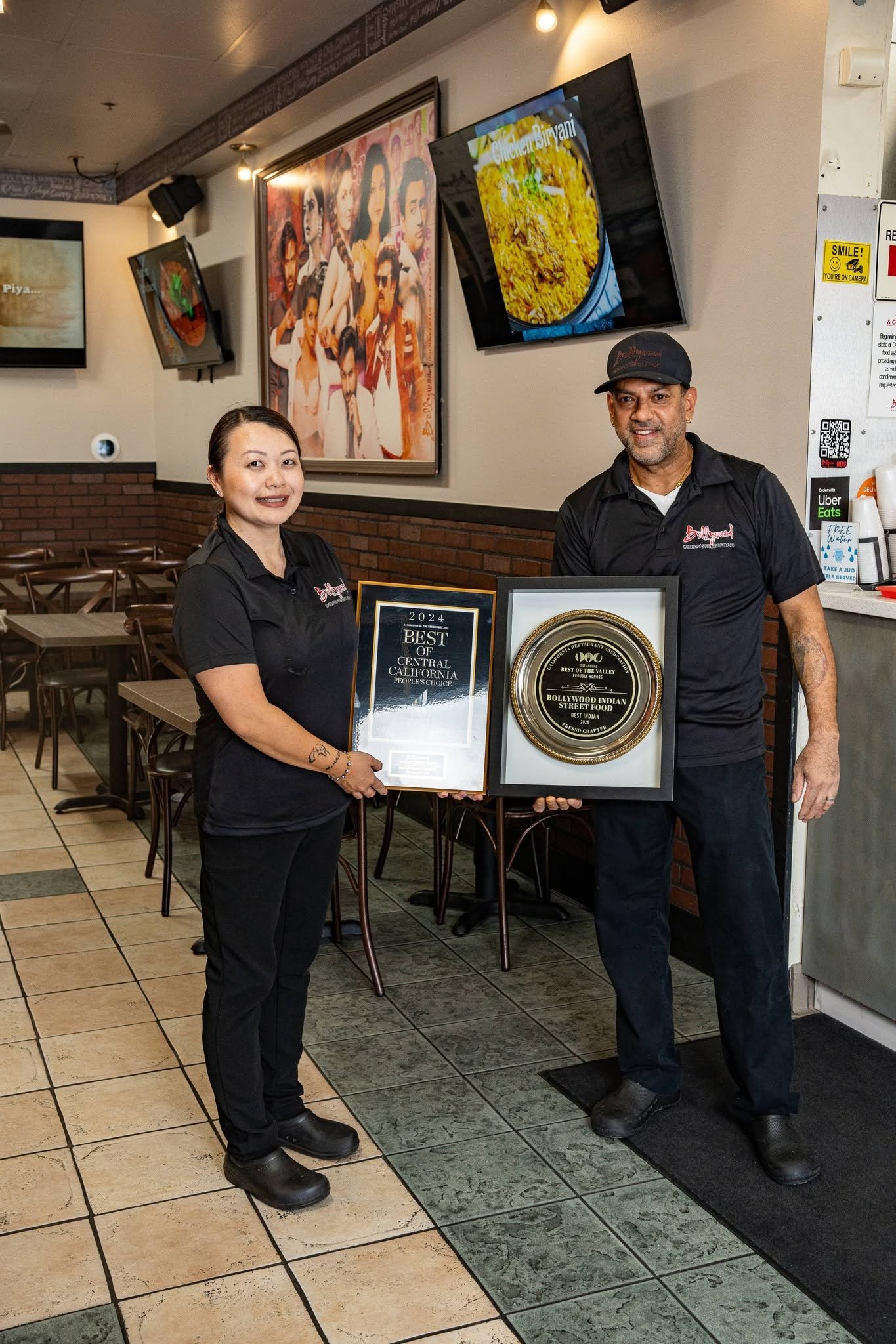 Two restaurant employees, a woman and a man, standing inside a casual dining restaurant. They are holding framed awards and medals. The woman is smiling and the man is wearing a cap. There are multiple television screens and artwork on the walls behind them. The restaurant has wooden chairs and tiled flooring.