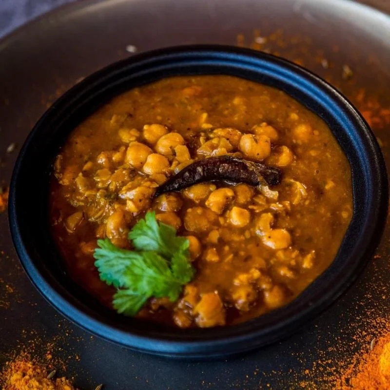 A bowl of chickpea curry garnished with cilantro and garnished with a dried chili, served in a black bowl.