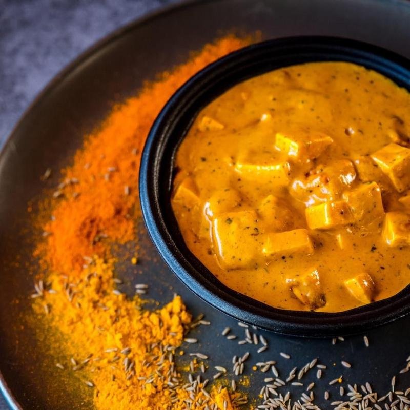 A bowl of creamy curry with chunks of tofu, served with a bed of rice and surrounded by saffron-colored spices.