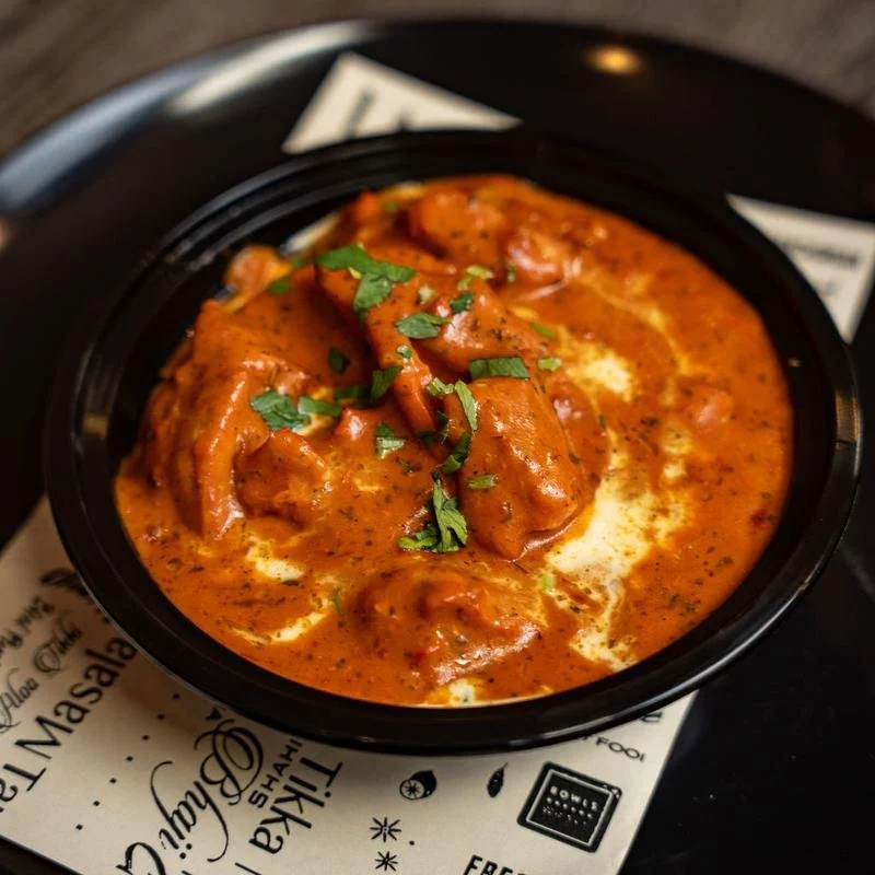 A black bowl of chicken curry garnished with chopped cilantro, served with a creamy sauce in a takeout container.