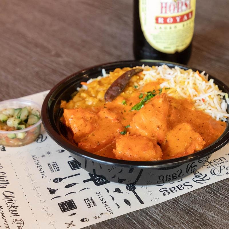 A bowl of Indian food with chicken curry, white rice, lentils, and a chili pepper, served on a paper with culinary words. A small side of cucumber salad and a bottle of beer are also visible.