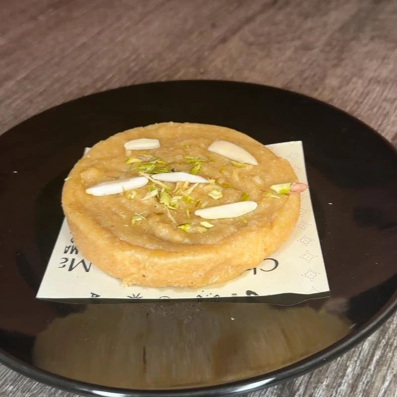 A round, fried Indian sweet dish topped with sliced almonds and pistachios, served on a white piece of paper on a black plate.